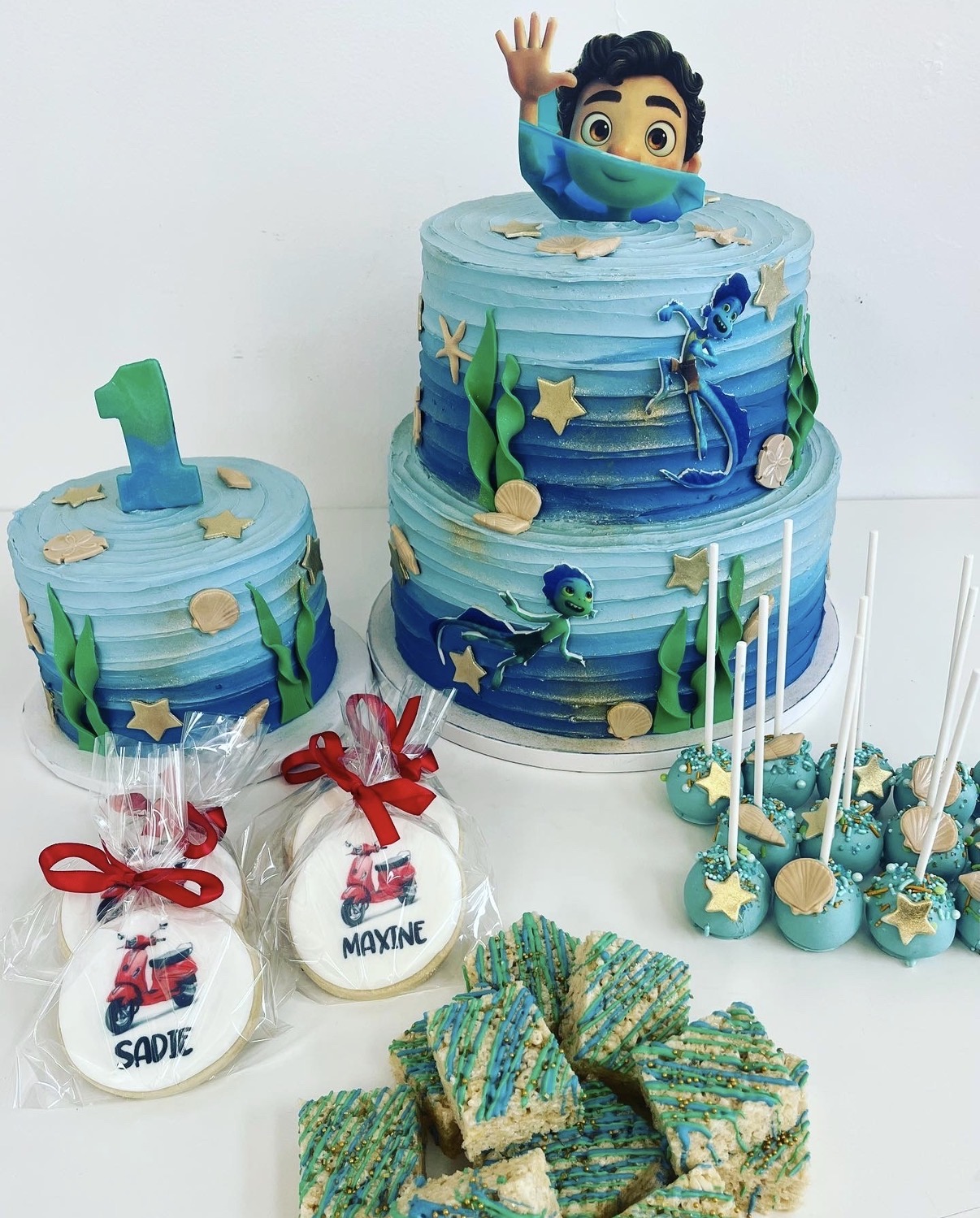 Two blue ocean-themed cakes with a character topper, surrounded by cake pops and decorated cookies, celebrating a child's first birthday.