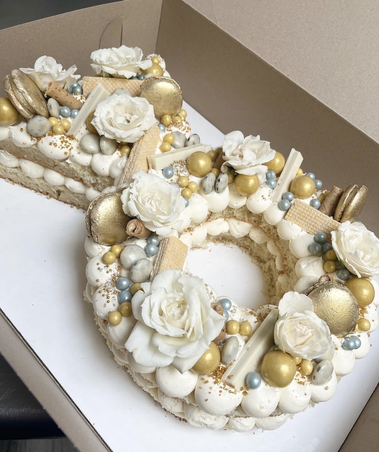 Decorated cake with white flowers, gold and silver balls, and cream-colored decorations inside a box.