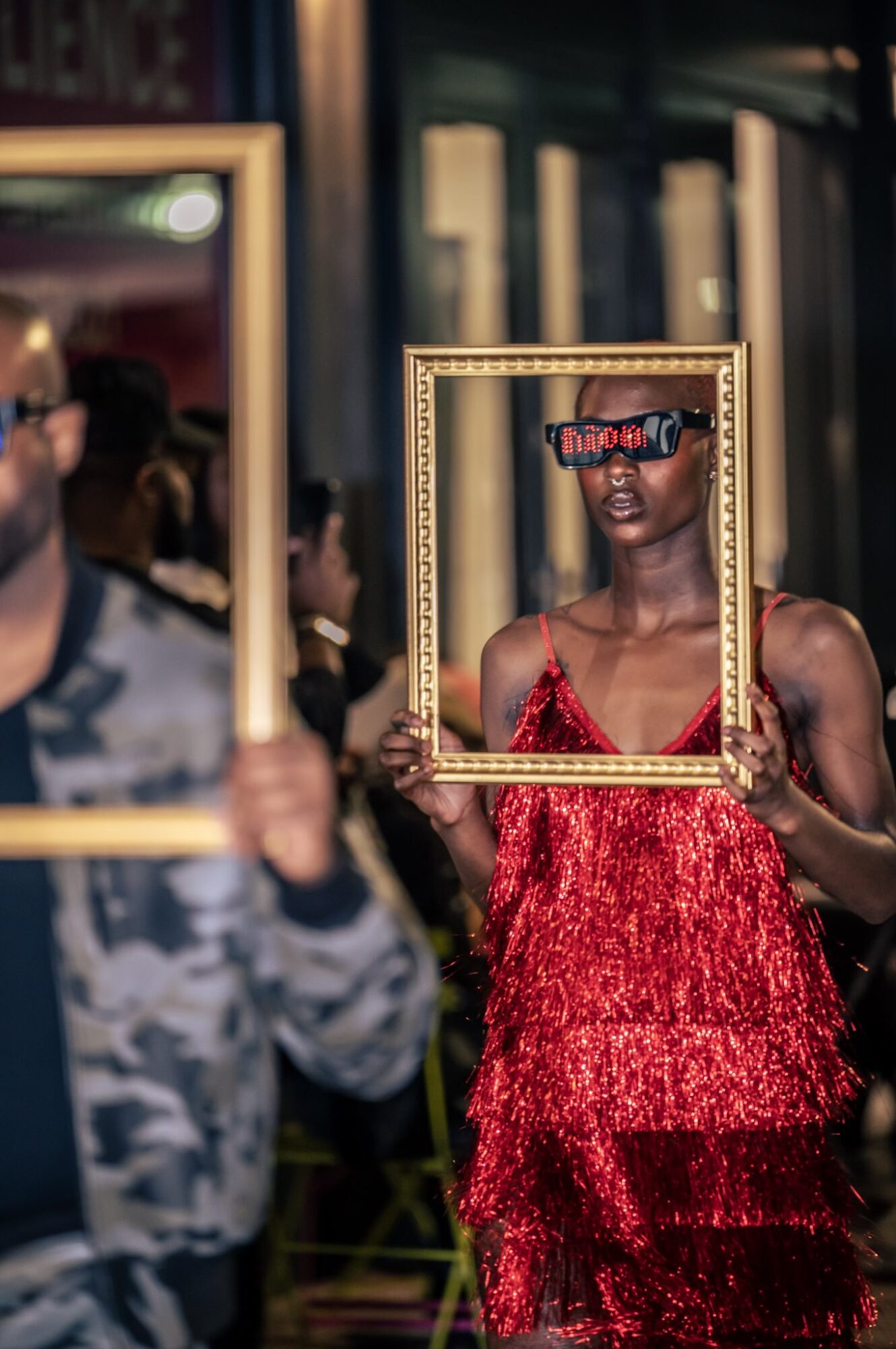 Woman in red dress holding a picture frame over her face, wearing sunglasses, in a dimly lit indoor setting.