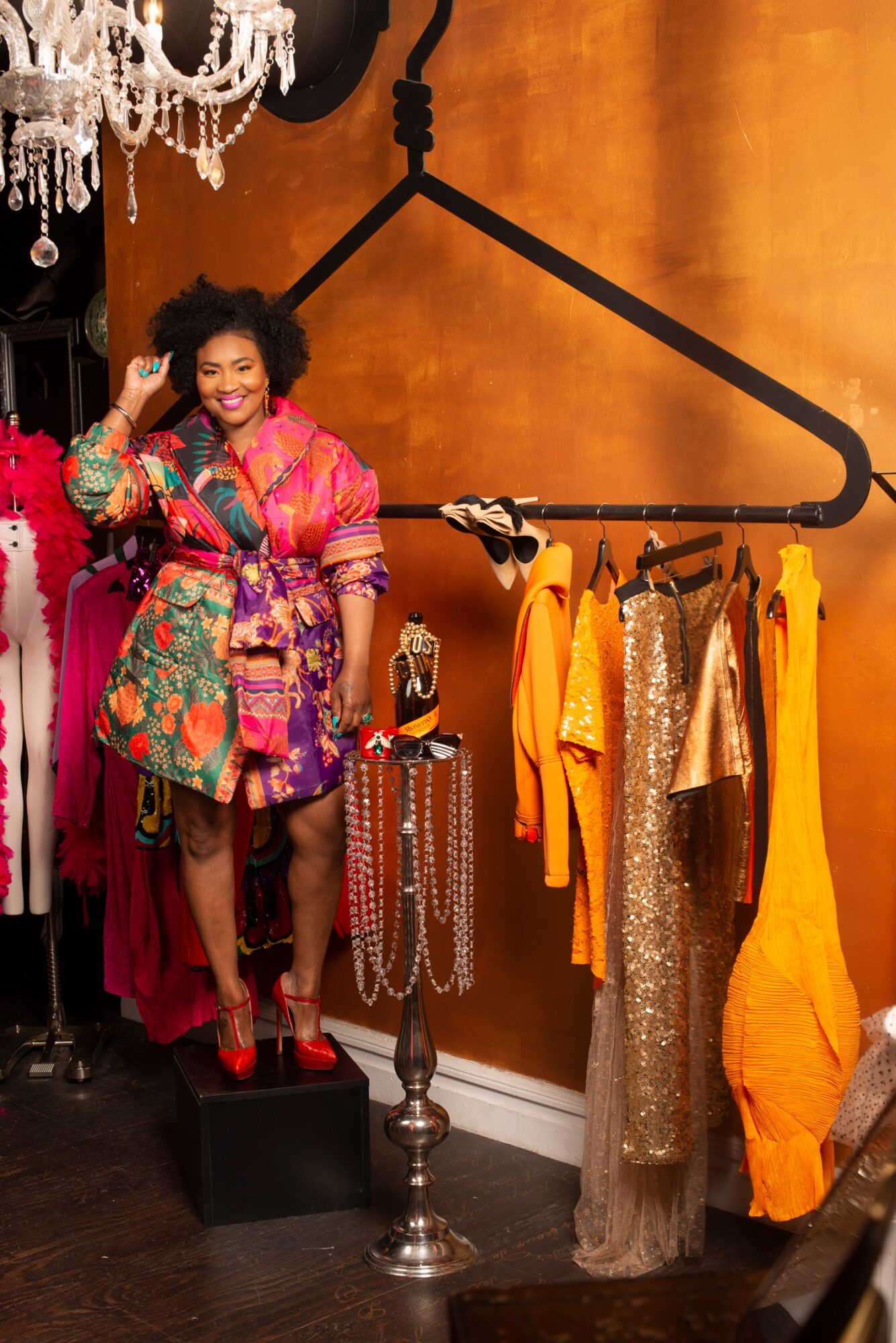 Woman with curly hair in colorful clothing standing on a platform, surrounded by clothing and accessories, in a boutique or dressing room.