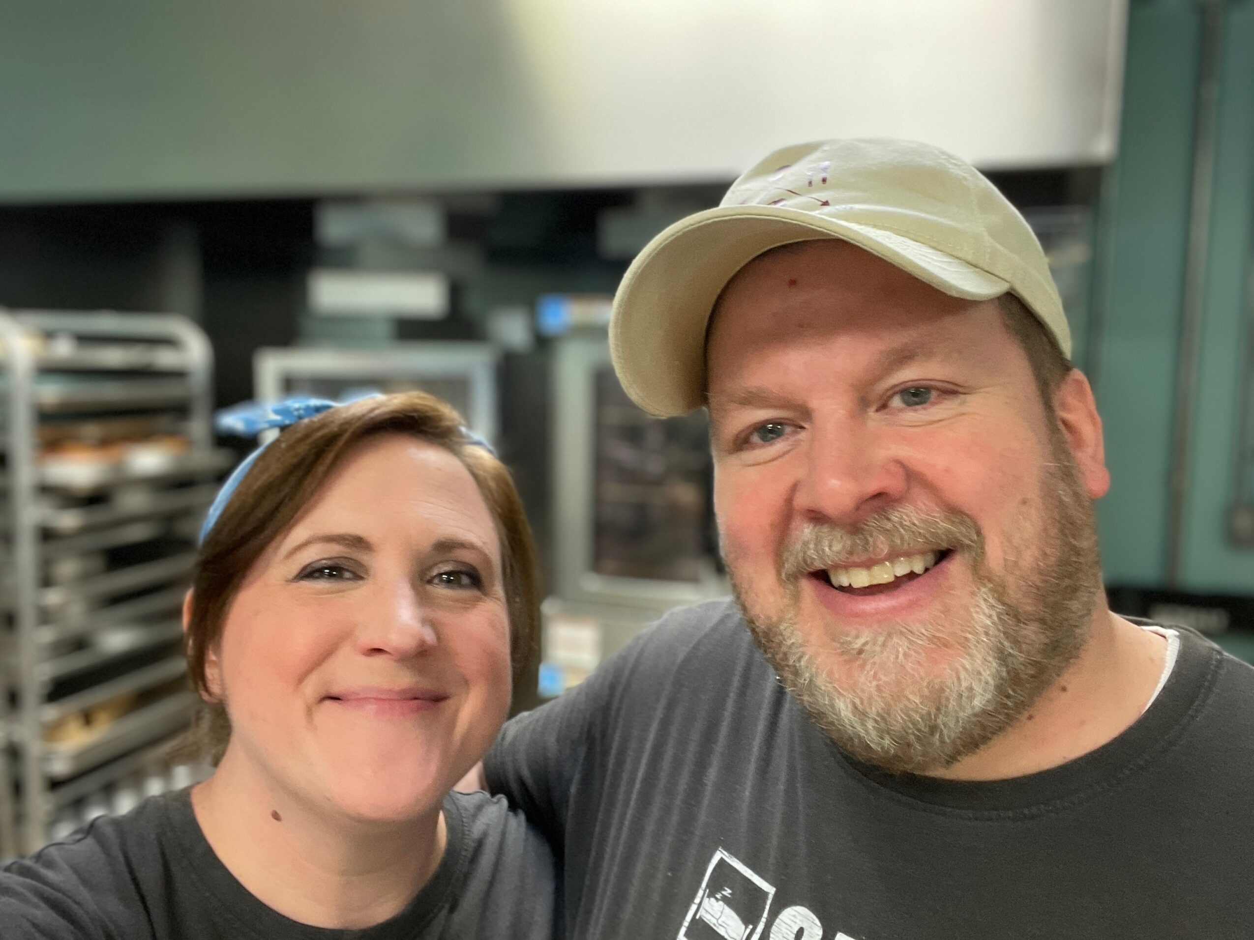 Two people smiling in a kitchen, one wearing a beige cap, the other with short brown hair, background with kitchen equipment.