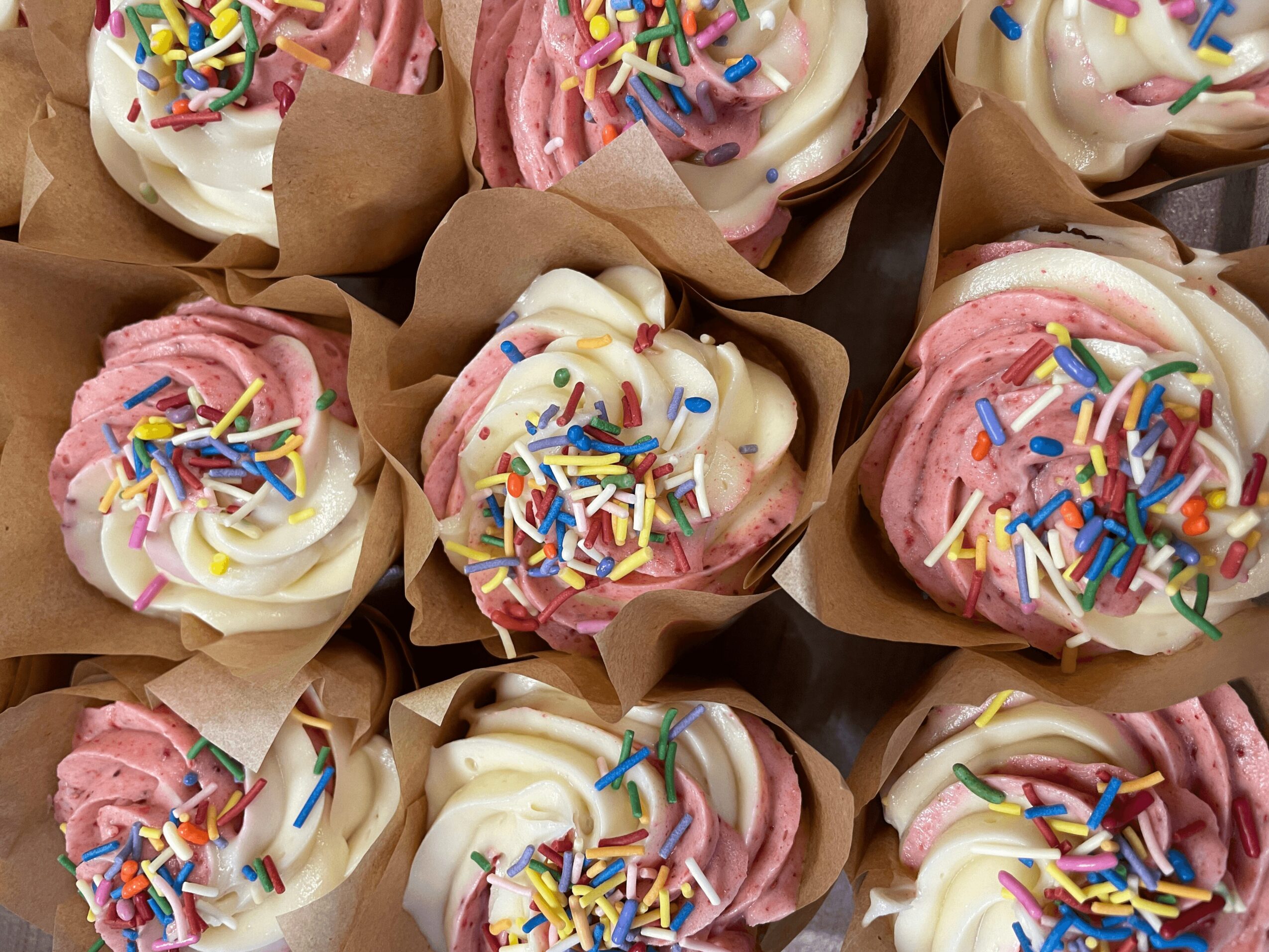 Cupcakes with white and pink frosting, colorful sprinkles, in brown paper wrappers.