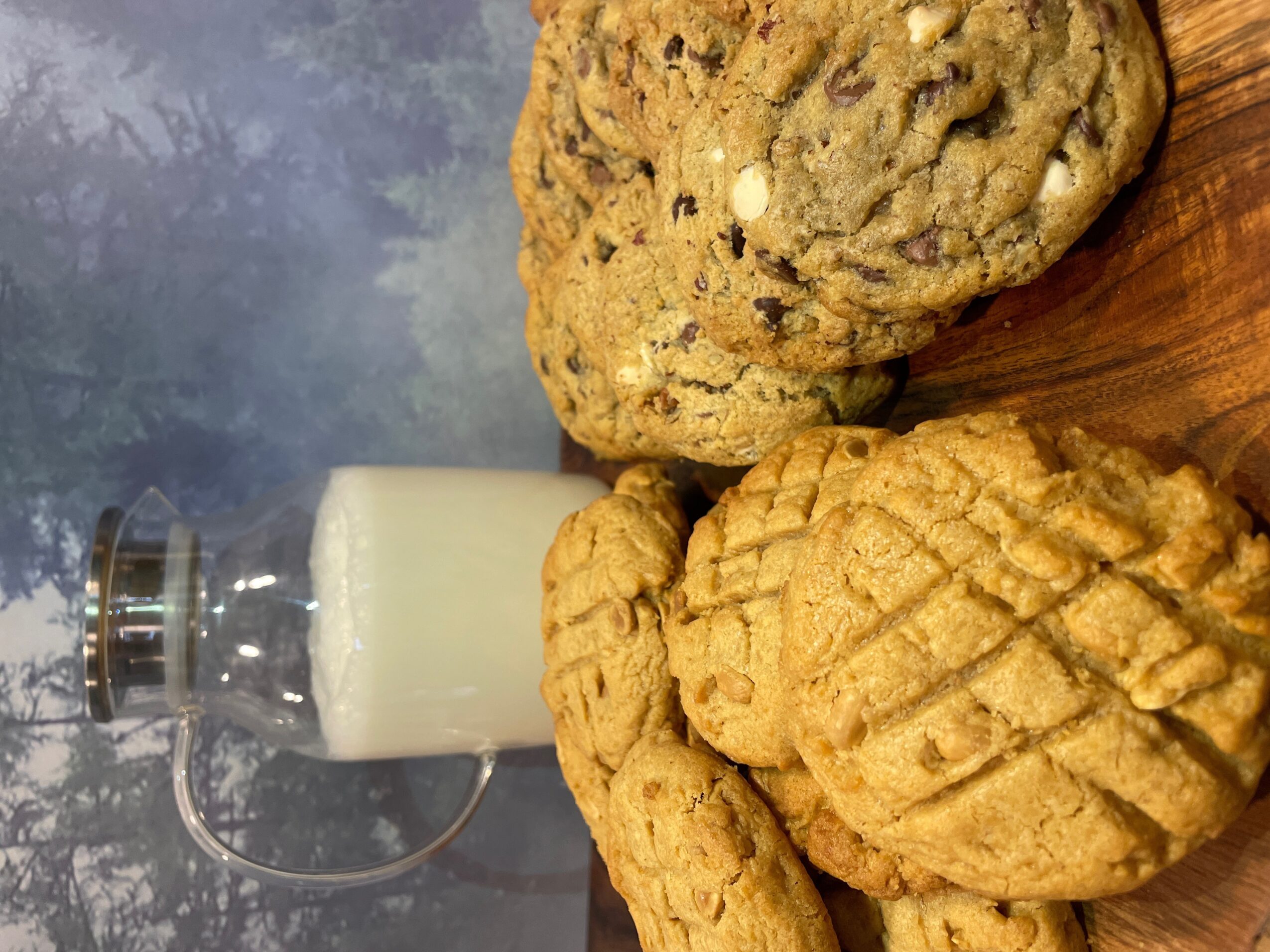 Three cookies and a glass of milk on a wooden surface, with a background of trees and sky.