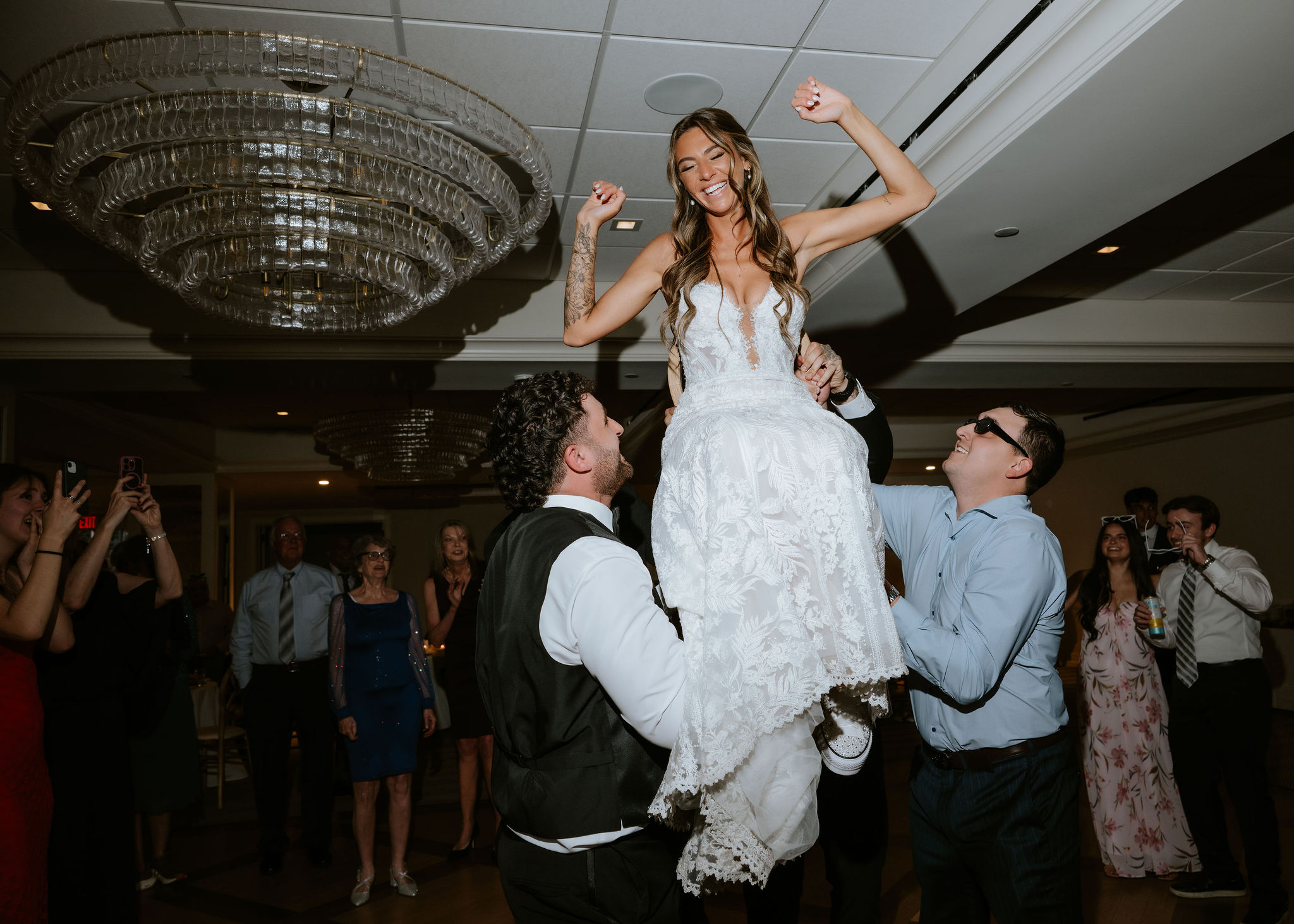 A woman in a wedding dress is lifted by two men at a celebration, with guests dancing in the background.
