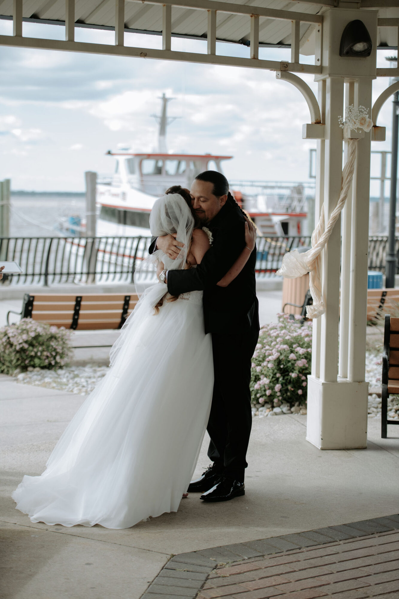 A bride and groom embrace under a pavilion near water with a boat in the background, wedding attire, and floral decorations.