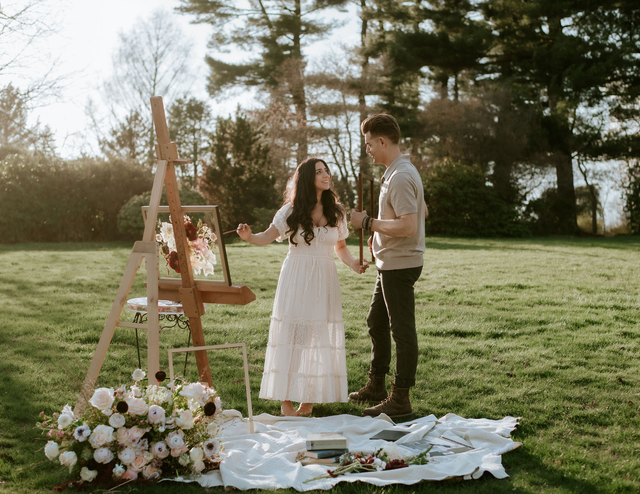 A couple stands outdoors near an easel with a painting, surrounded by flowers and art supplies on a white cloth.