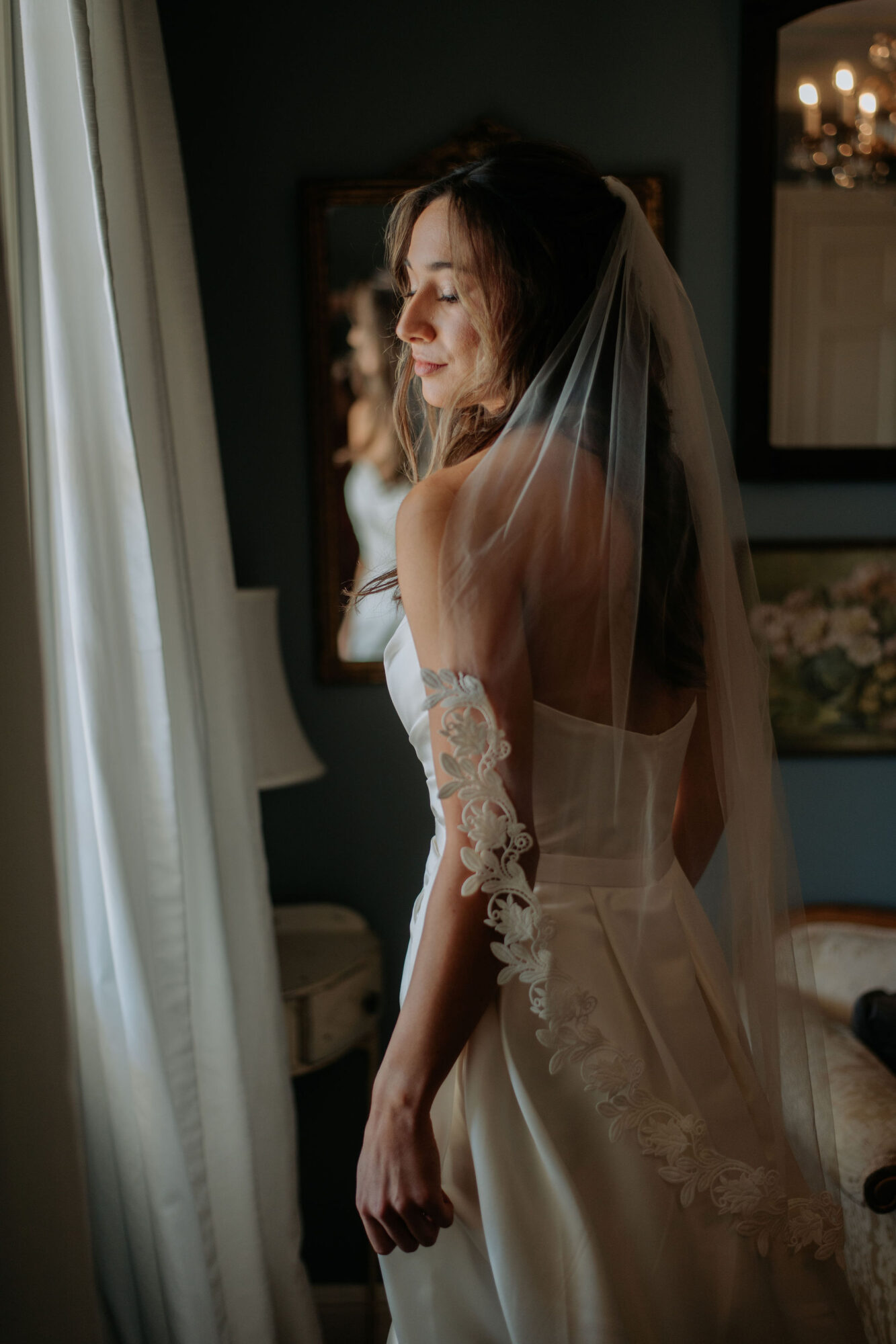 Bride with veil looking out window, soft light illuminating her face and dress, in a dimly lit room.