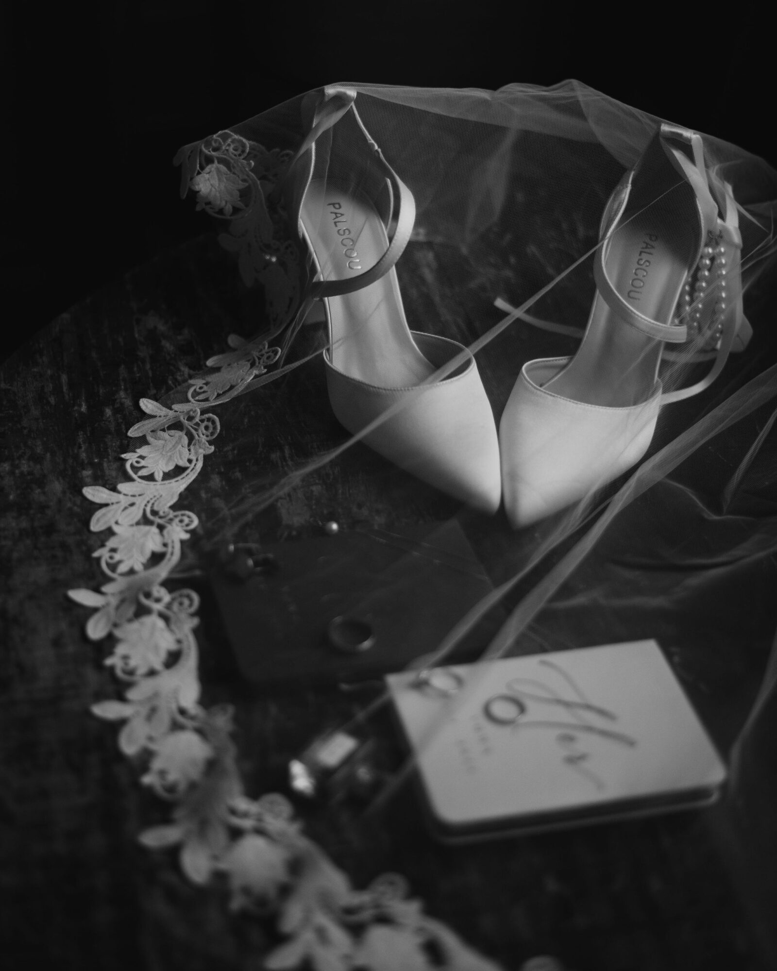 Pair of white high-heeled shoes with lace veil, floral decoration, and a card on a dark surface.