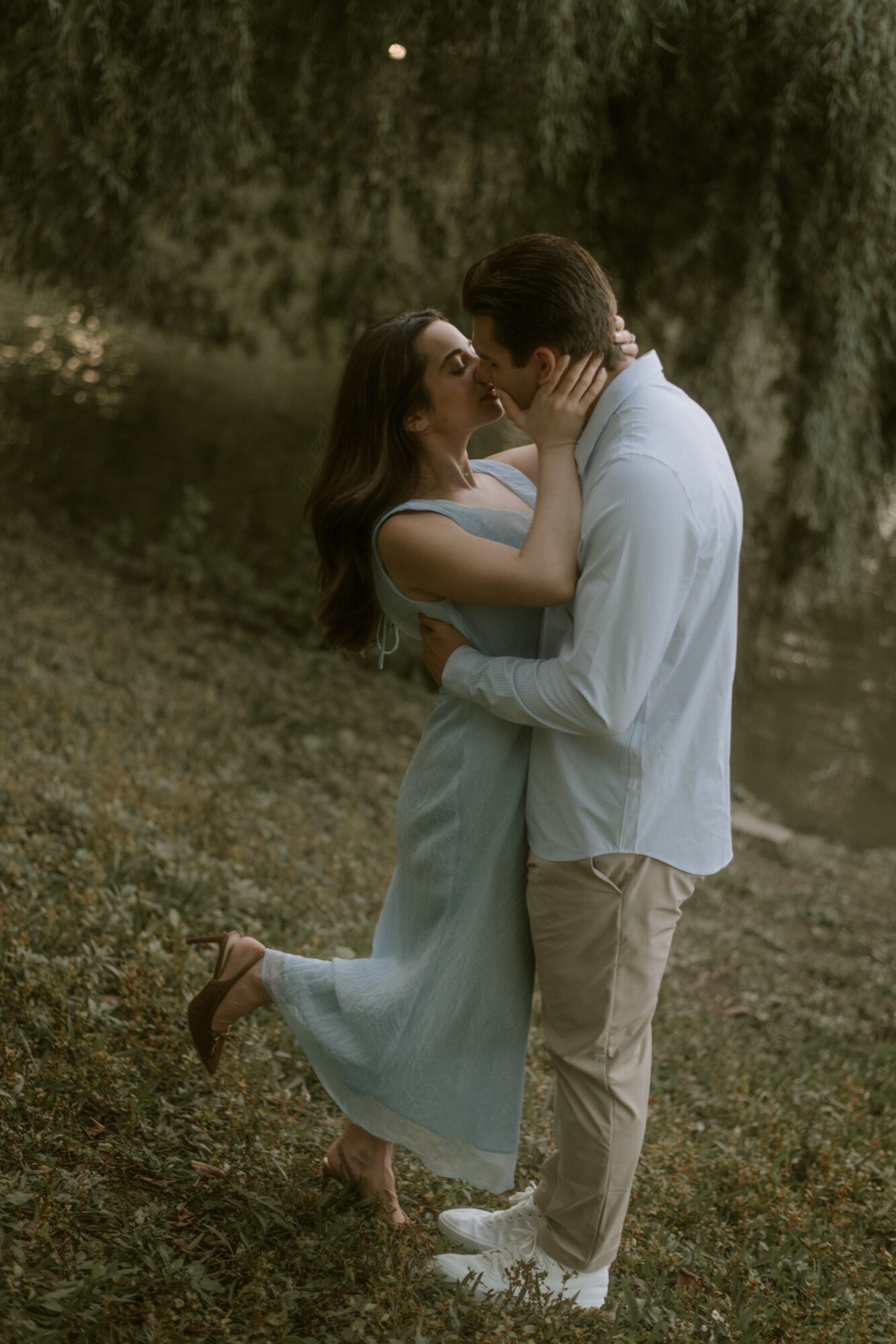 A couple stands close, touching foreheads and holding hands outdoors with trees and grass in the background.