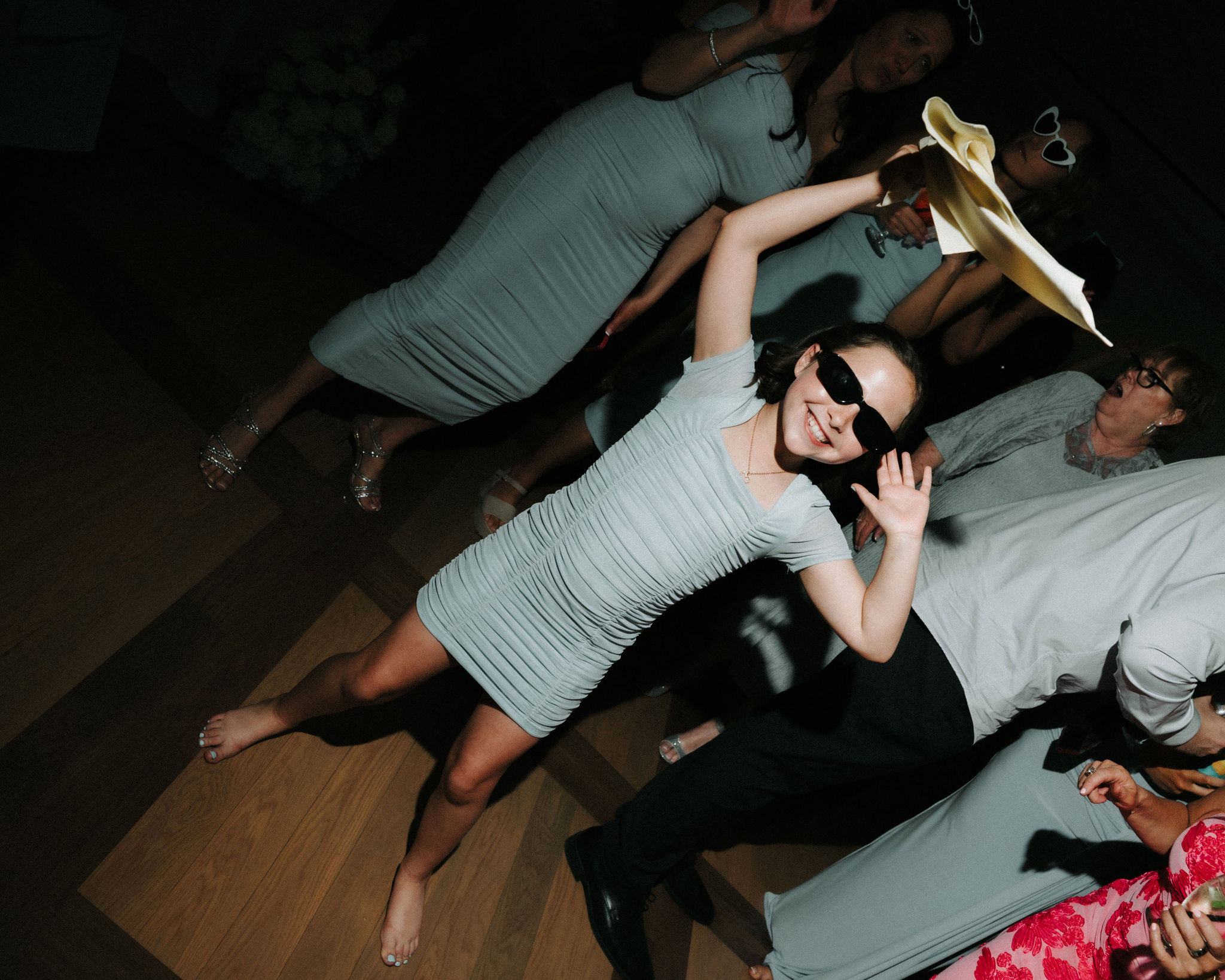 Woman in a light-colored dress and sunglasses dancing with a hat, smiling, in a party setting.