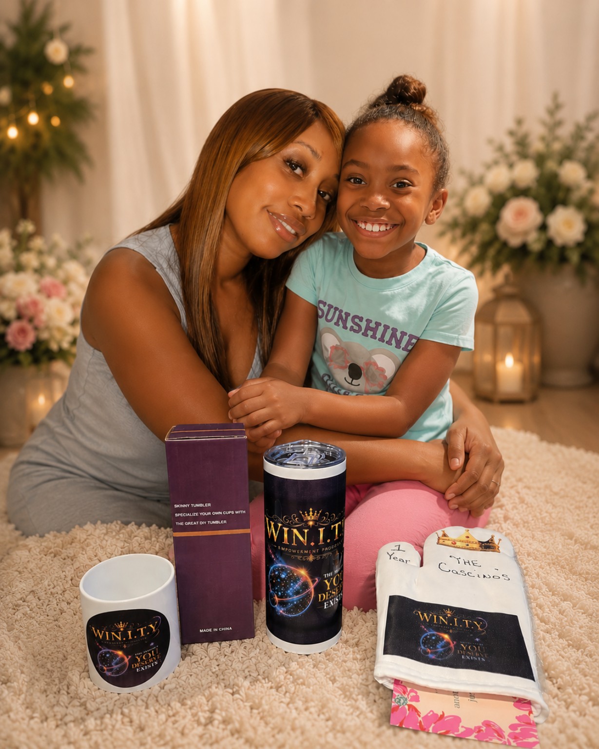 Woman and girl smiling, sitting on a carpet with gift items, flowers, and candles in background.
