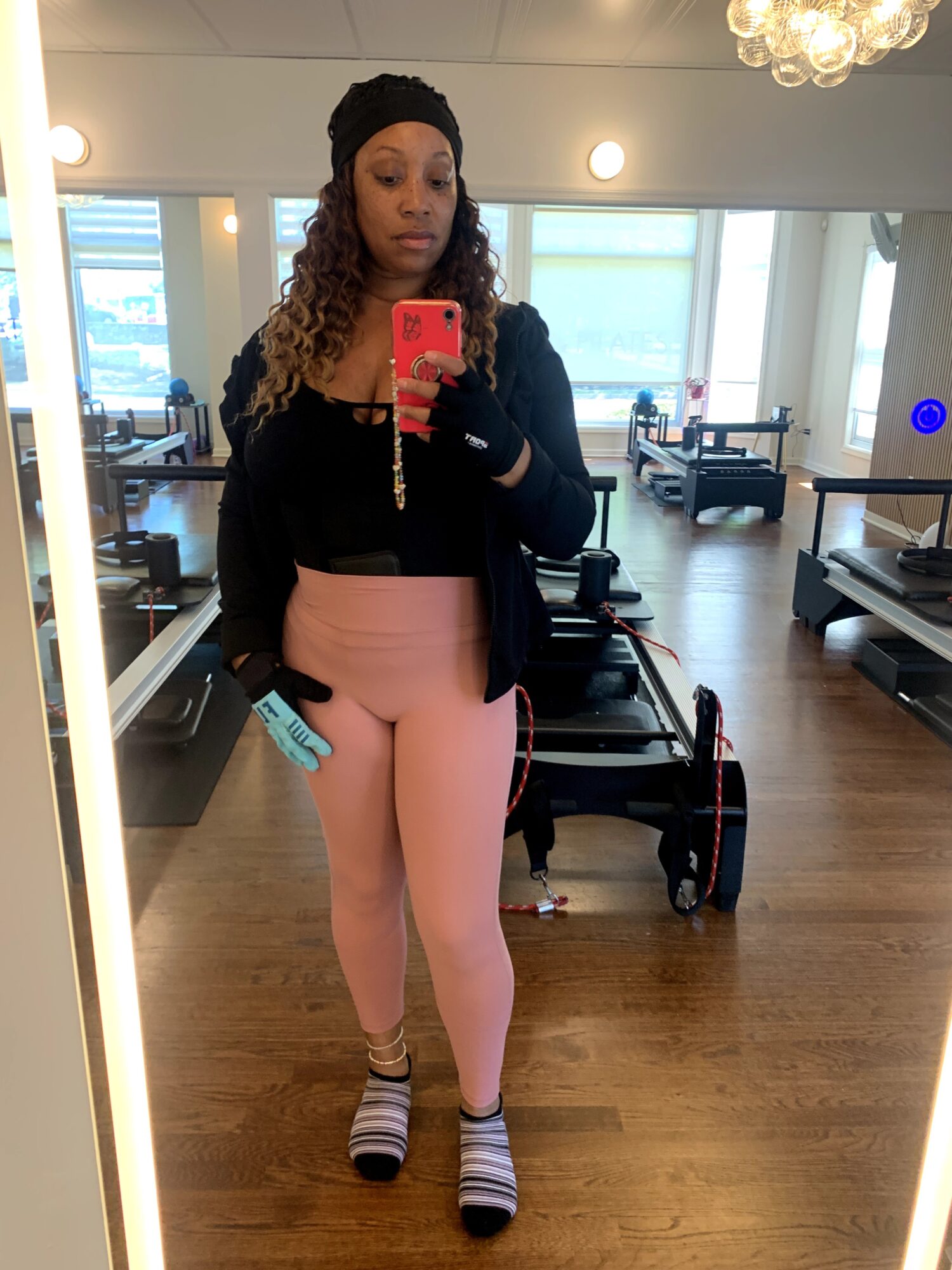 Woman taking a mirror selfie in a room with wooden floors, large windows, and modern furniture, wearing a black jacket, pink pants, and striped socks.