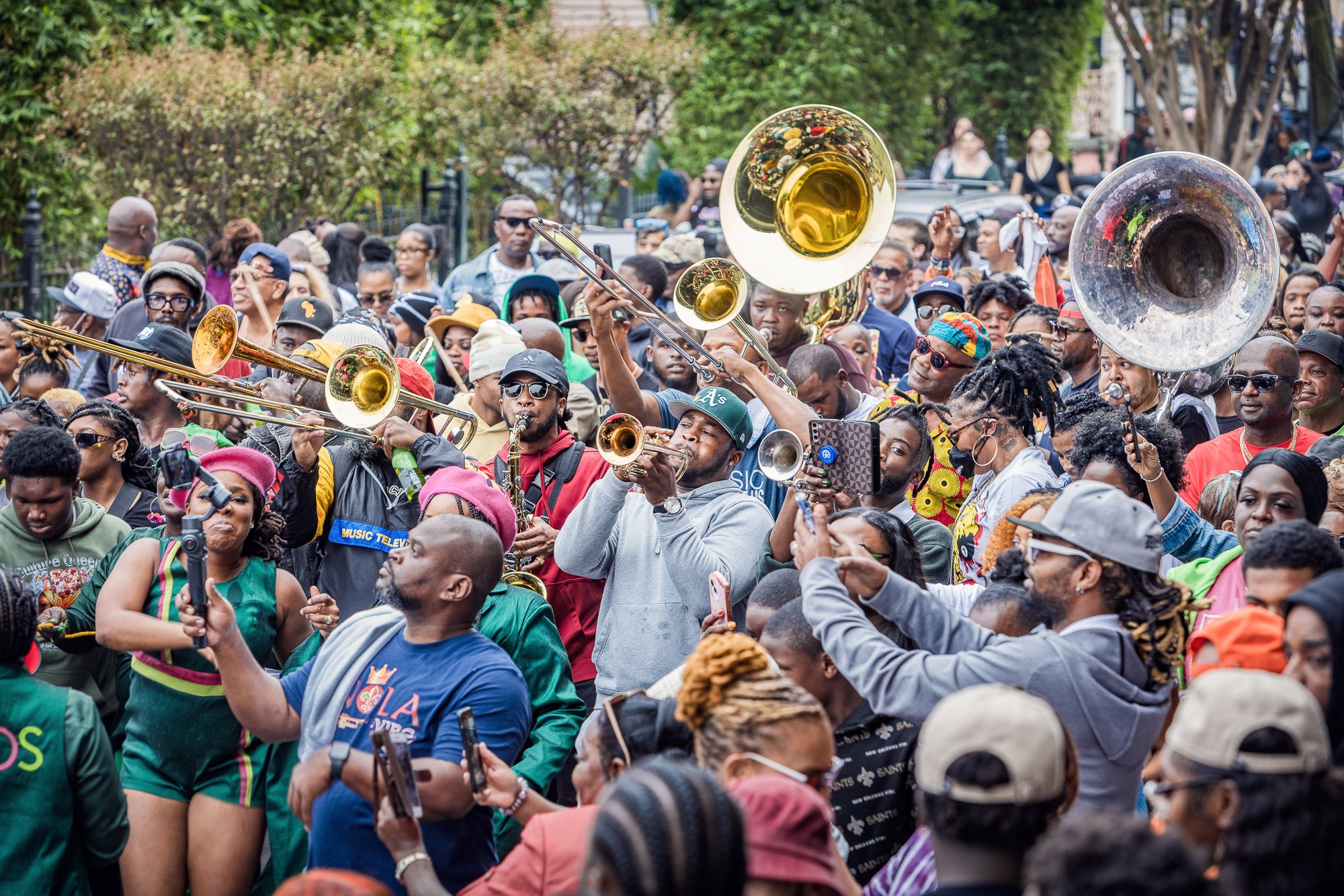 Crowd of people playing brass instruments outdoors, some wearing hats and sunglasses, with trees and greenery in background.