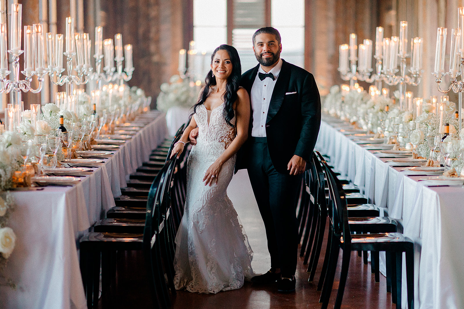A bride and groom stand together in a decorated banquet hall with long tables and chandeliers, smiling at the camera.