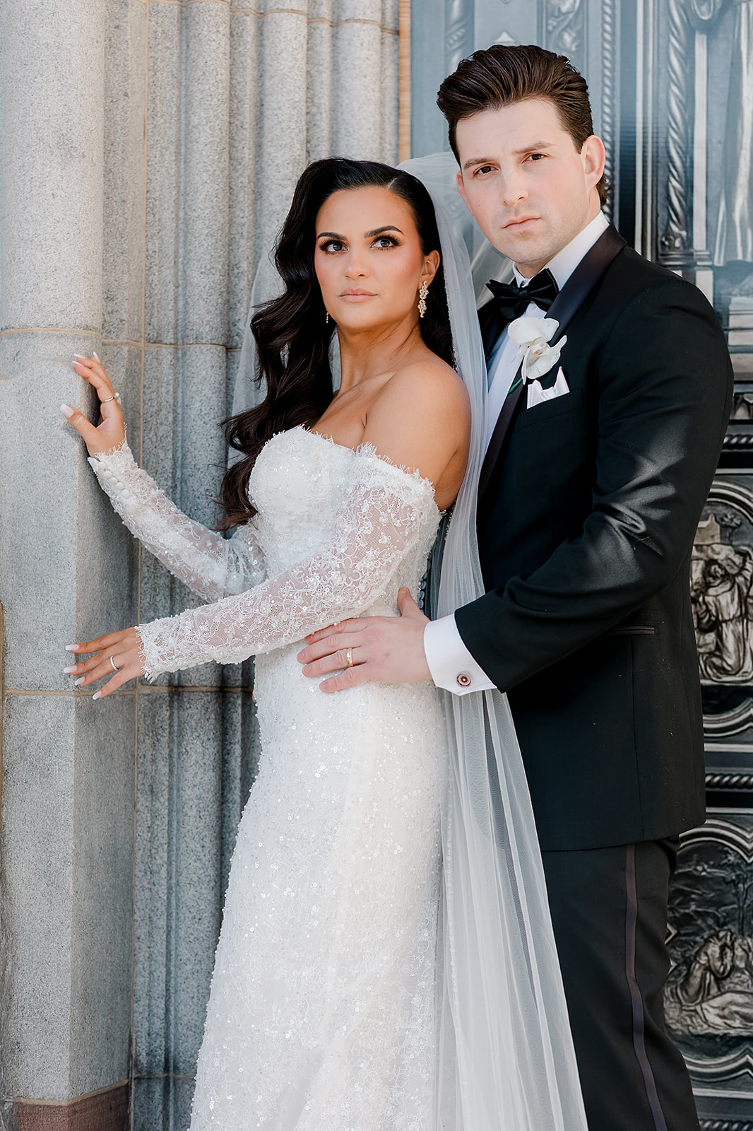 A bride and groom pose together, standing close, with the bride in a white gown and the groom in a tuxedo.