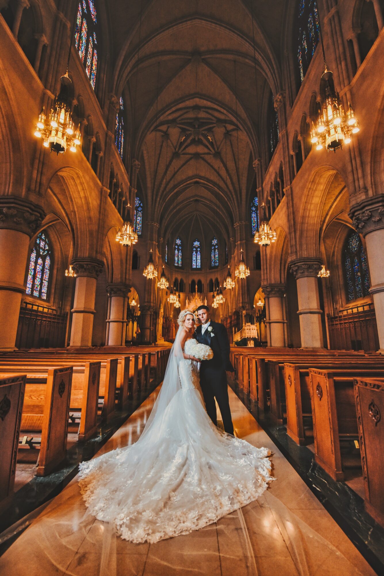 Bride and groom standing in a church aisle with high vaulted ceilings and ornate lighting fixtures.