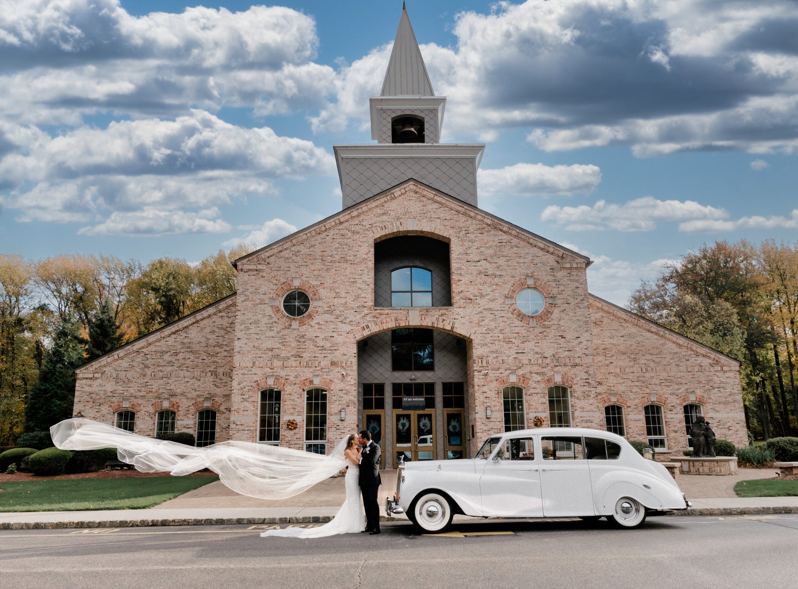 Bride and groom stand beside a vintage white car in front of a brick church with a steeple, under a cloudy sky.