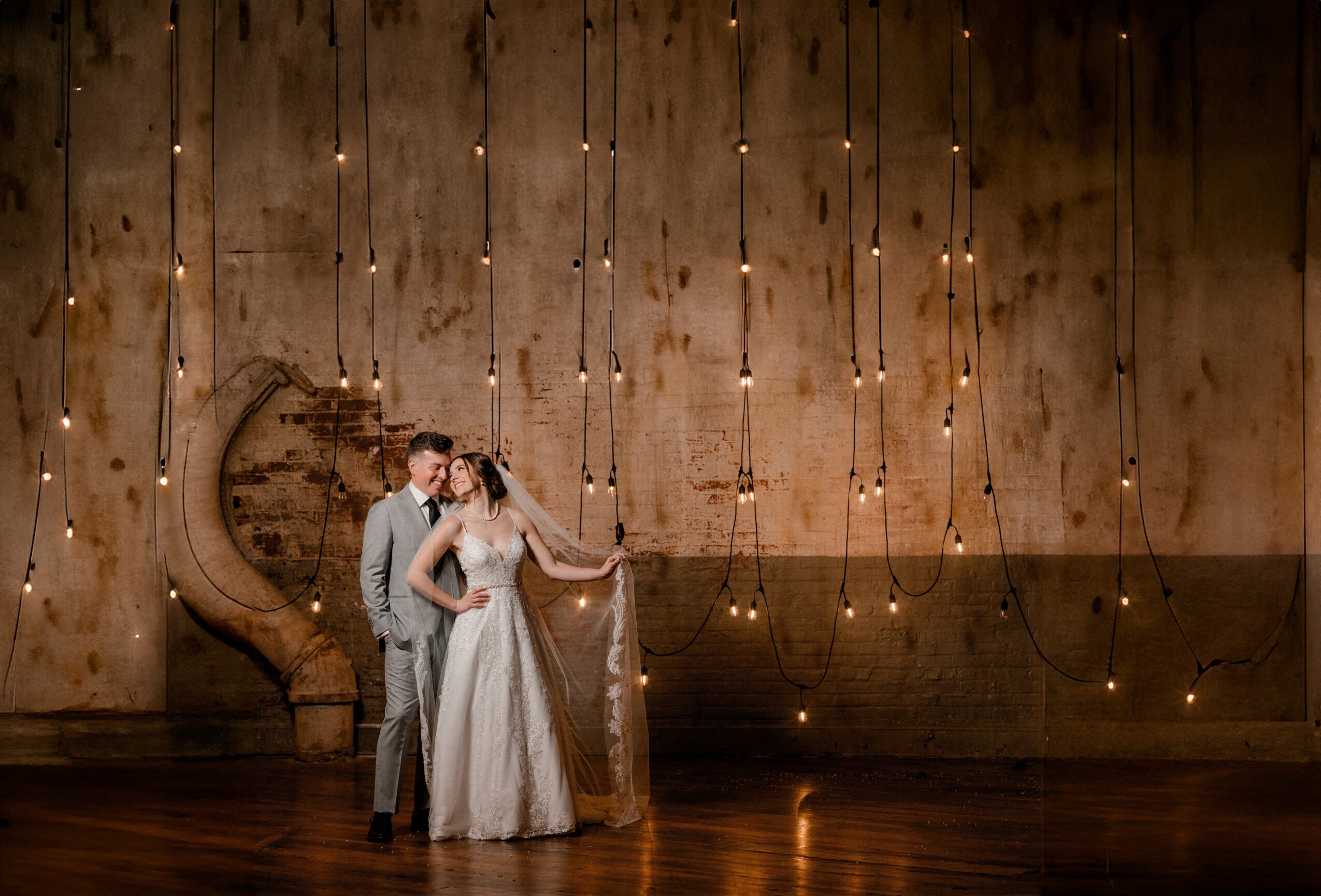 A bride and groom stand together on a wooden floor, with a woman, in front of a decorated wall with hanging lights.