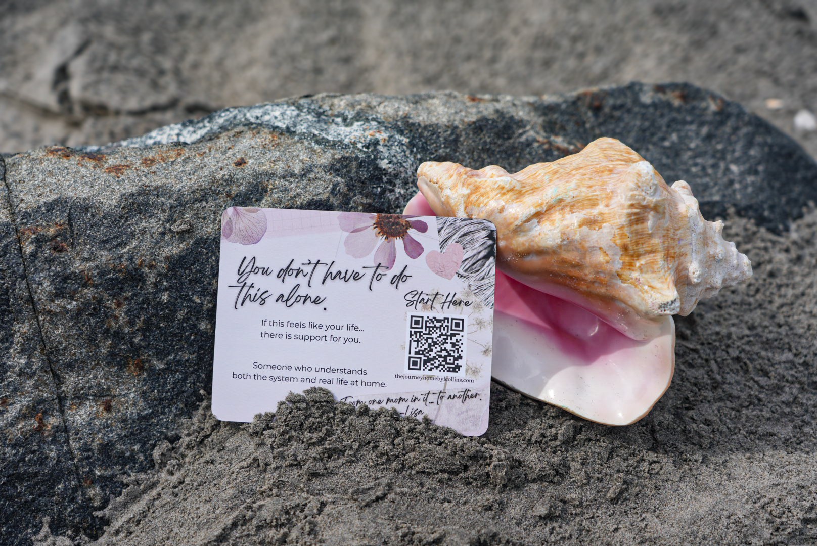 A seashell and a note card on rocks, with a QR code and handwritten message, outdoors.