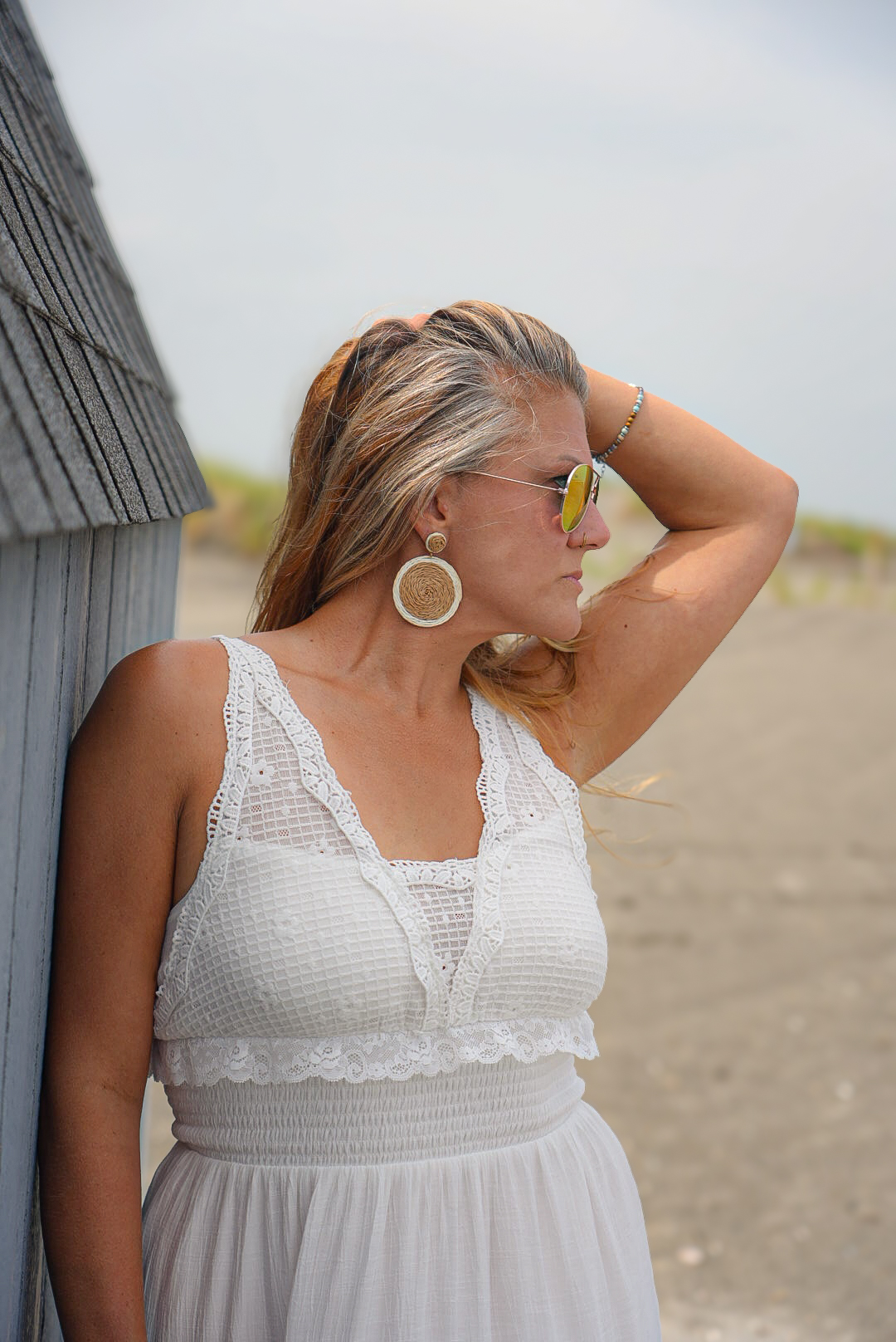 Woman with sunglasses and large earrings, wearing a white dress, standing outdoors with a cloudy sky and sandy background.