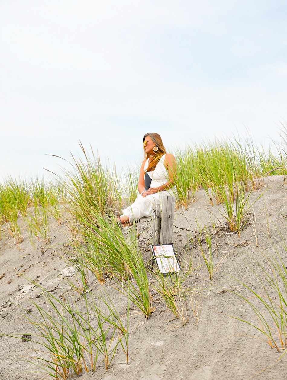Woman standing on sandy beach with grass, looking to the side, sky in background.