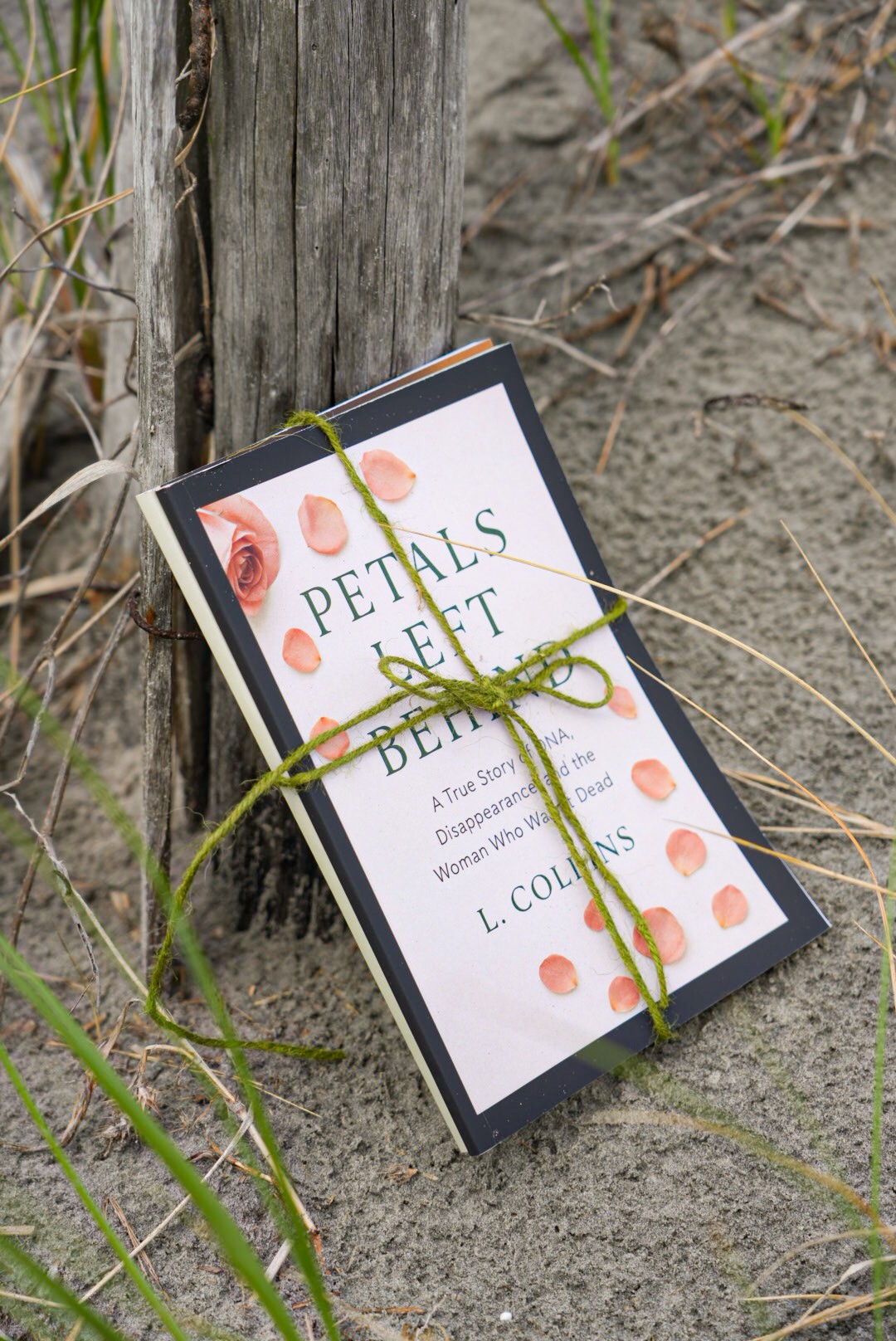 Book titled 'Petals & Sweet' tied with a green string, resting on sandy ground near a wooden post, surrounded by grass.