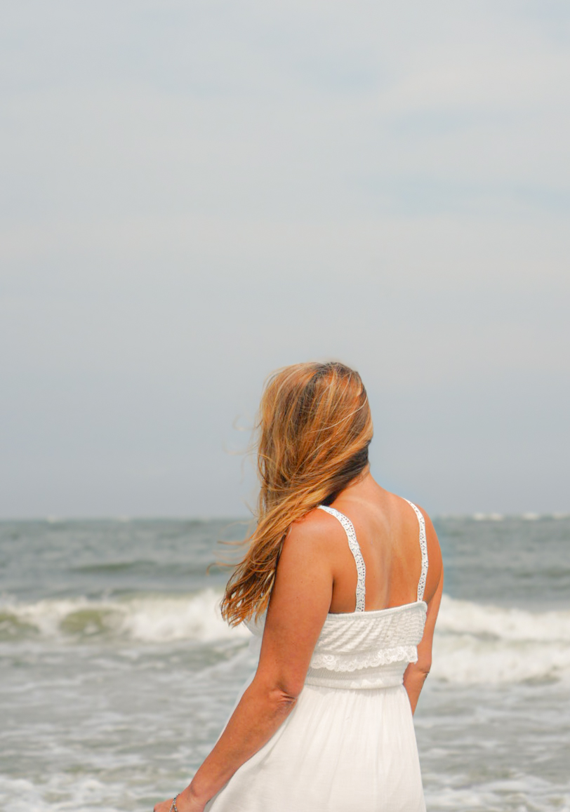 Woman with long hair in a white dress standing on the beach facing the ocean, waves in the background.