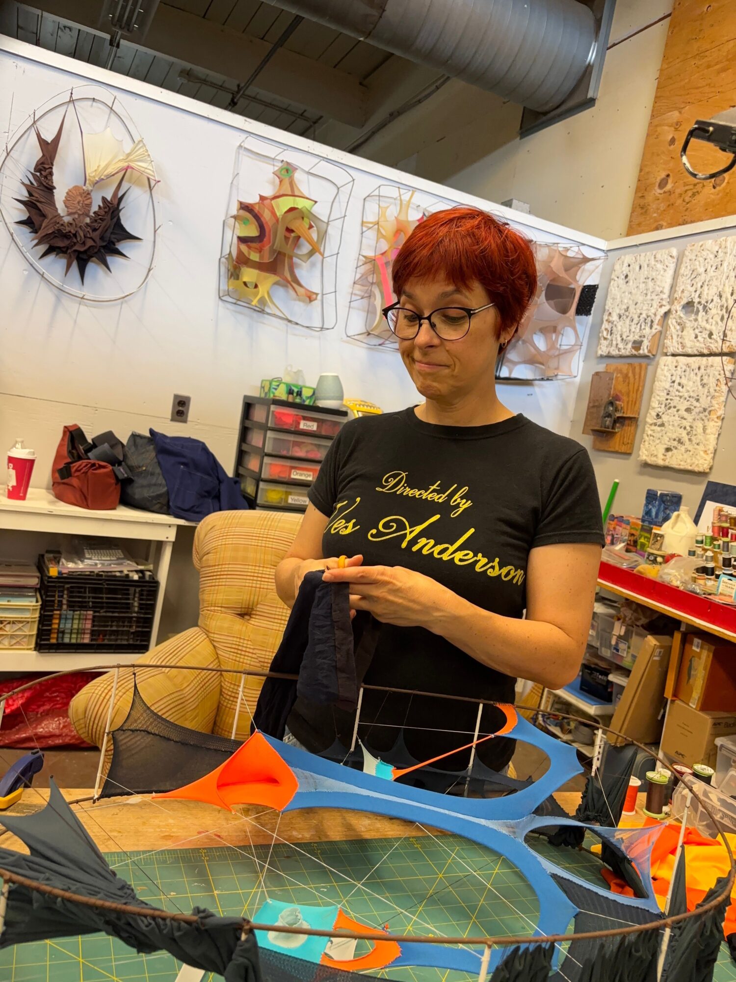 Woman with red hair and glasses working at a cluttered desk in a room with art supplies and posters.