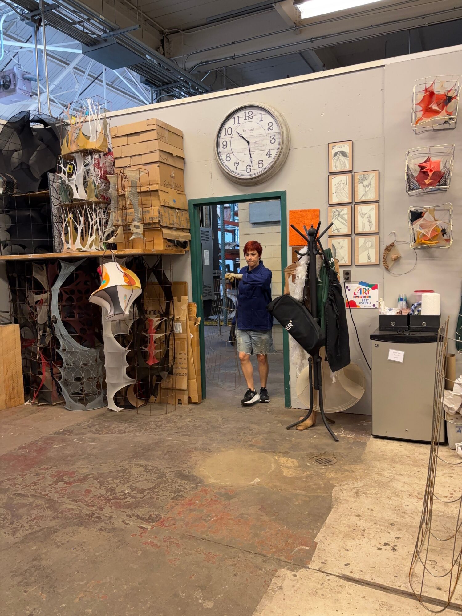 Person standing in a room with a clock, boxes, and various items on shelves, holding a bag and a coat.