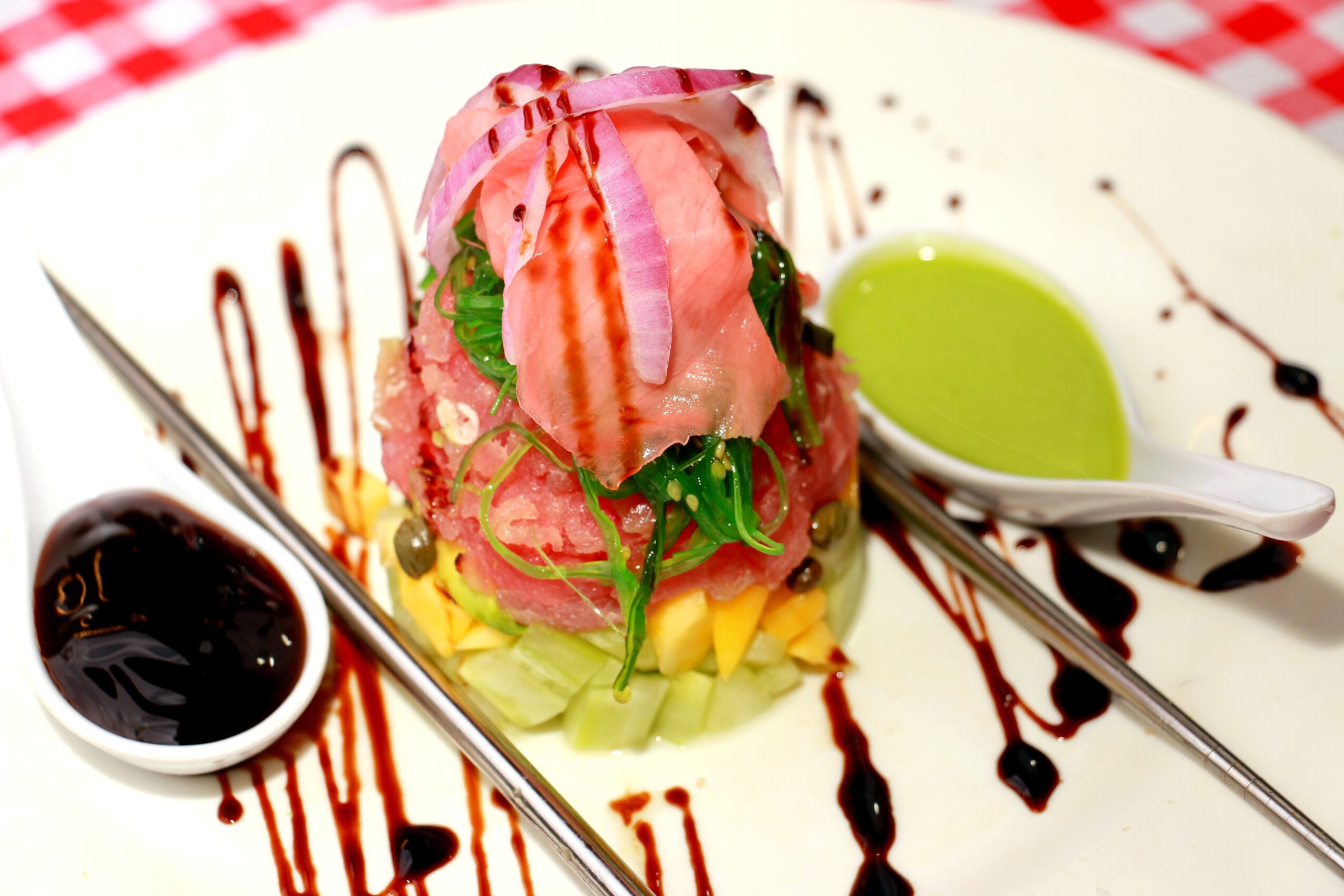 A dessert with pink ice cream, fruit, and green sauce, decorated with chocolate drizzle and served with a spoon and fork.
