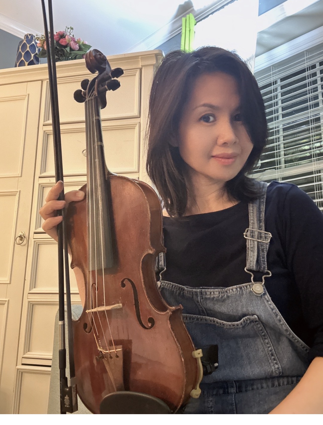 Young woman with shoulder-length dark hair holding a violin indoors, smiling at the camera, wearing a black top and denim overalls.