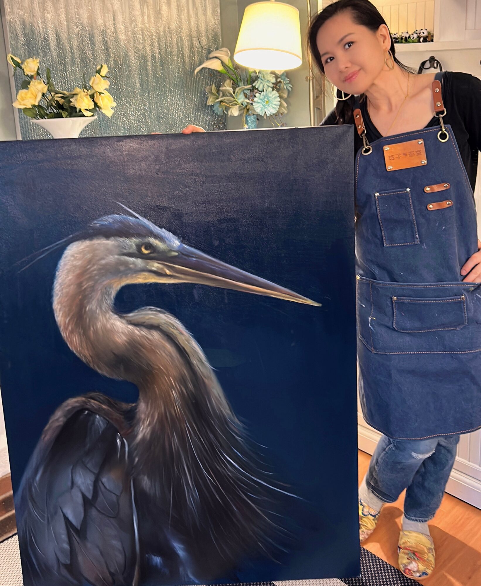 Woman standing next to a painting of a heron, indoors with flowers and a lamp in the background.