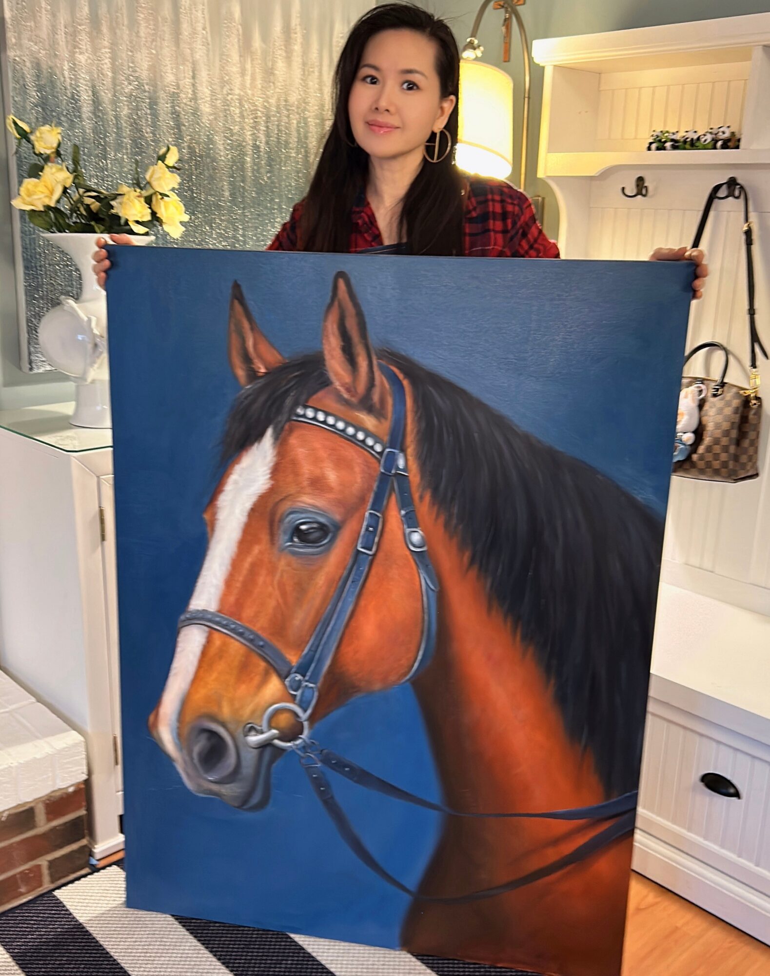 Woman holding a painting of a brown horse with a blue background in a room with white furniture and yellow flowers.