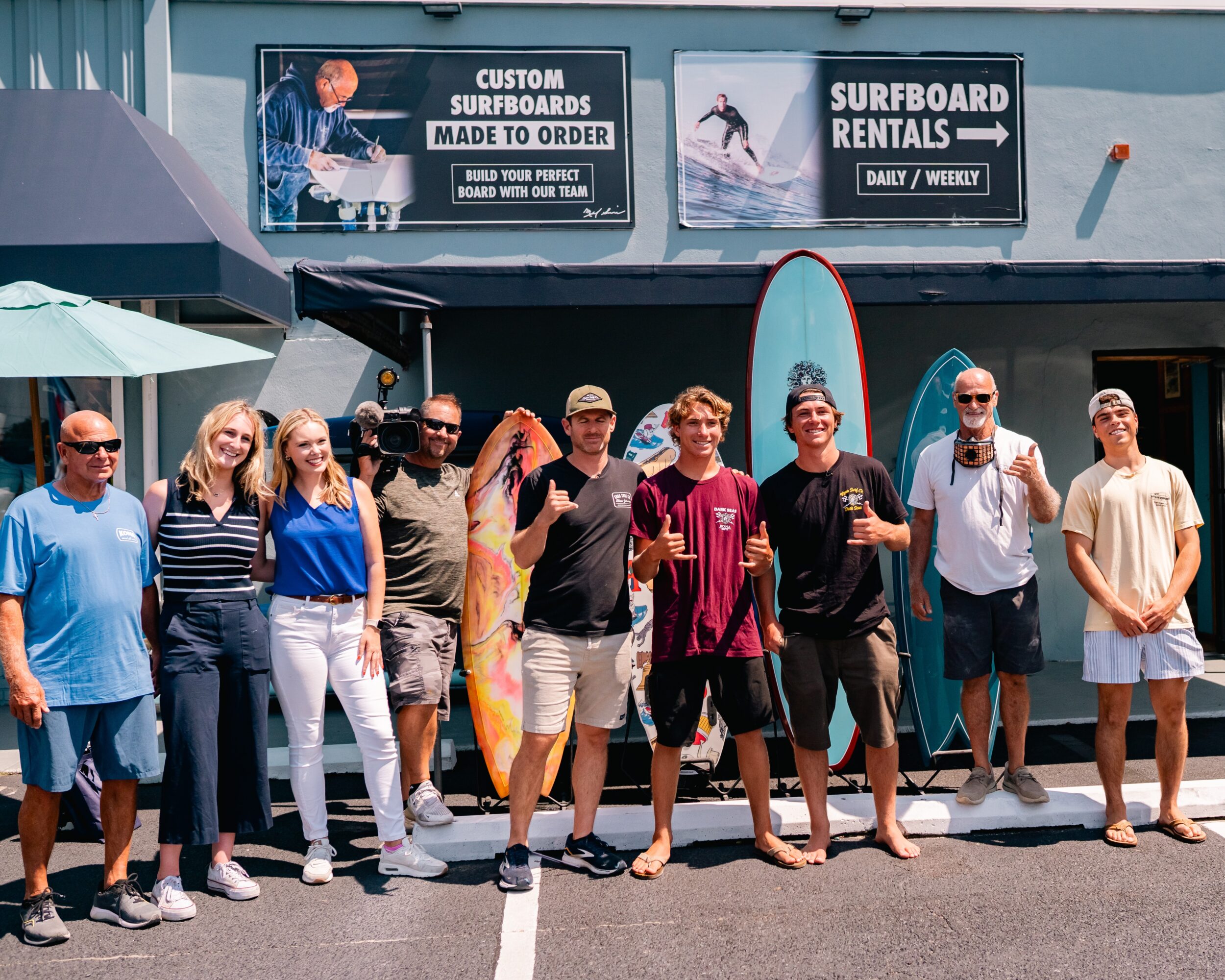 Group of nine people standing outside surf shop, some holding surfboards, smiling, with signs for surfboard rentals and custom surfboards behind them.