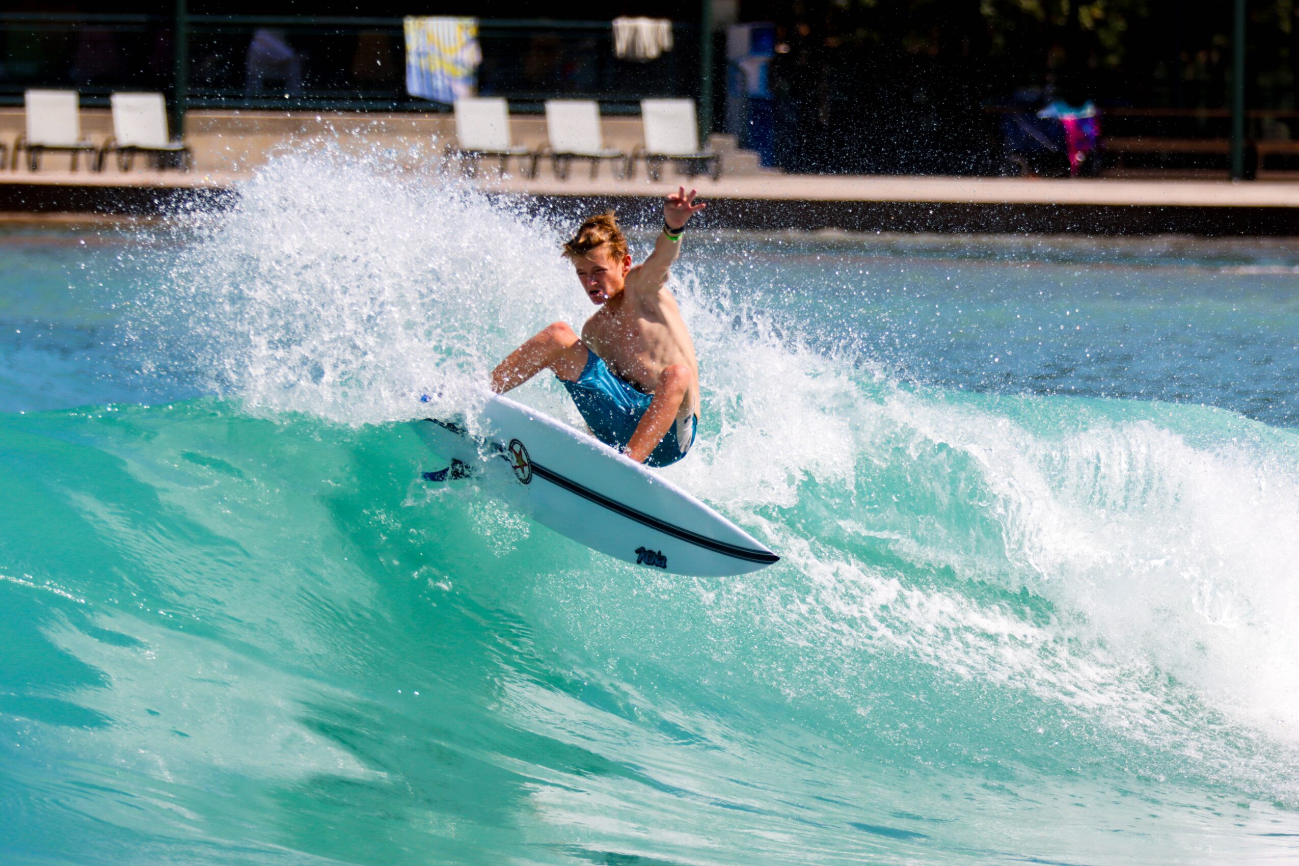 Person surfing on a wave in a pool, with splash and water spray, background shows poolside chairs and people.