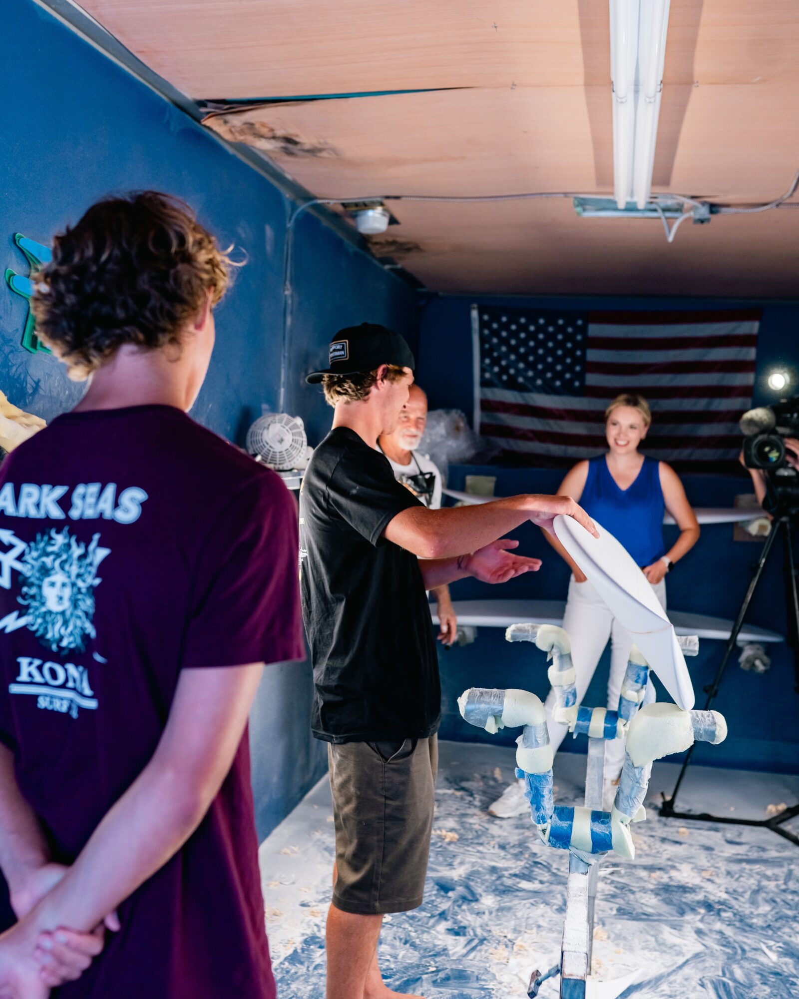 Young person operating a robot with a woman observing, in a room with an American flag and snow-like floor, ceiling damaged.