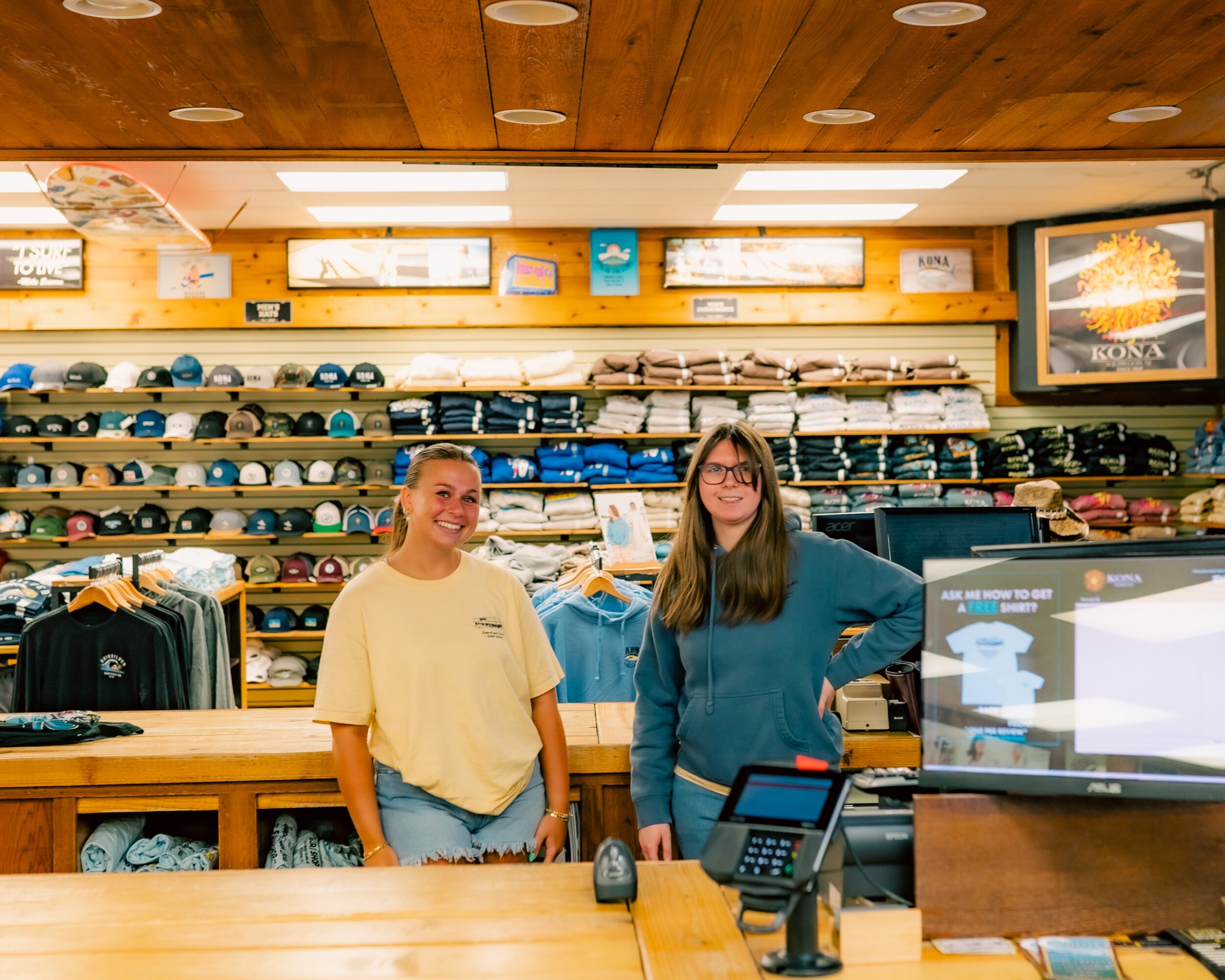 Two women stand behind a store counter with shelves of hats and clothing in the background.