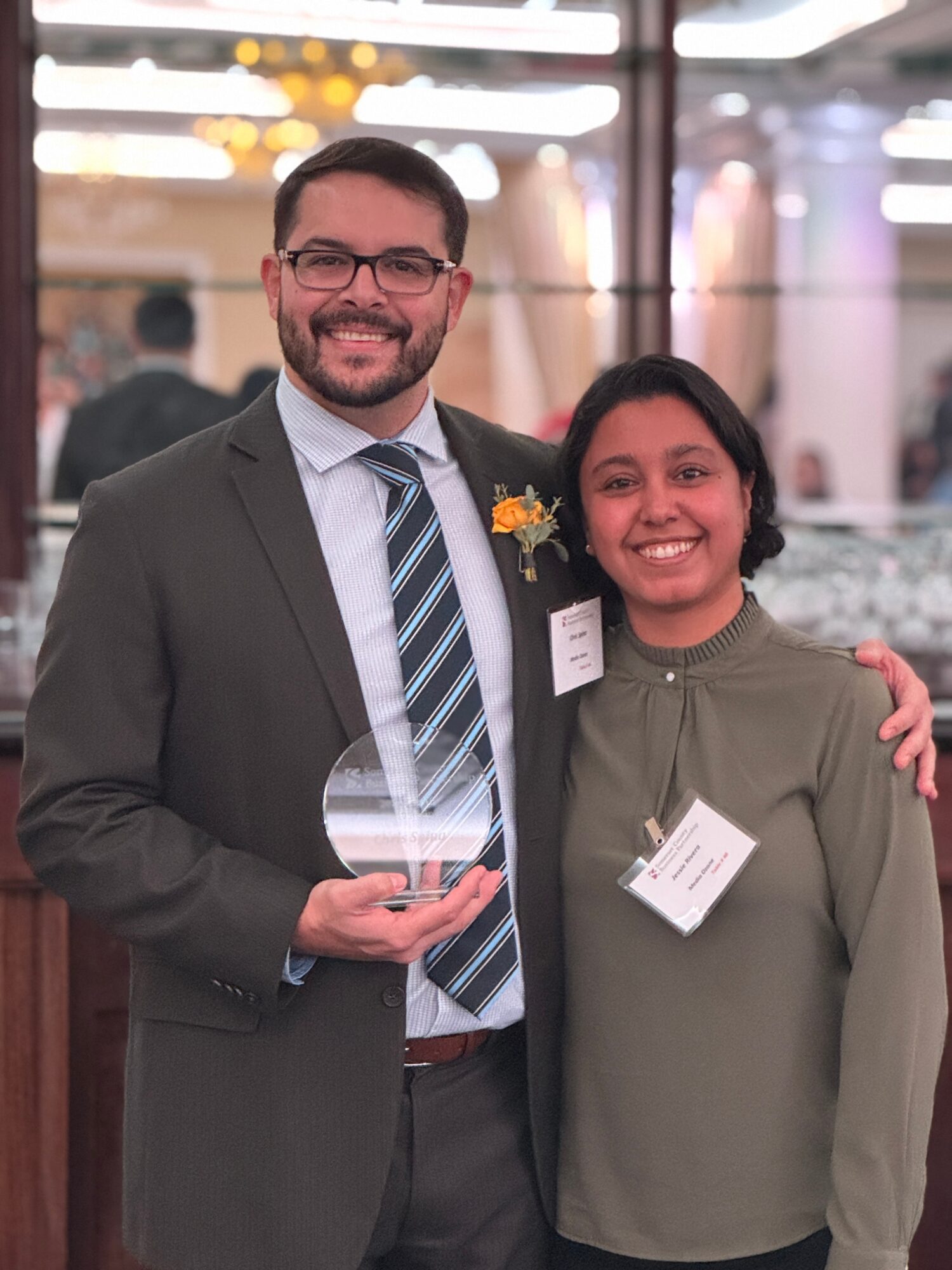 Two people smiling, one holding an award, in a formal setting with blurred background lights.