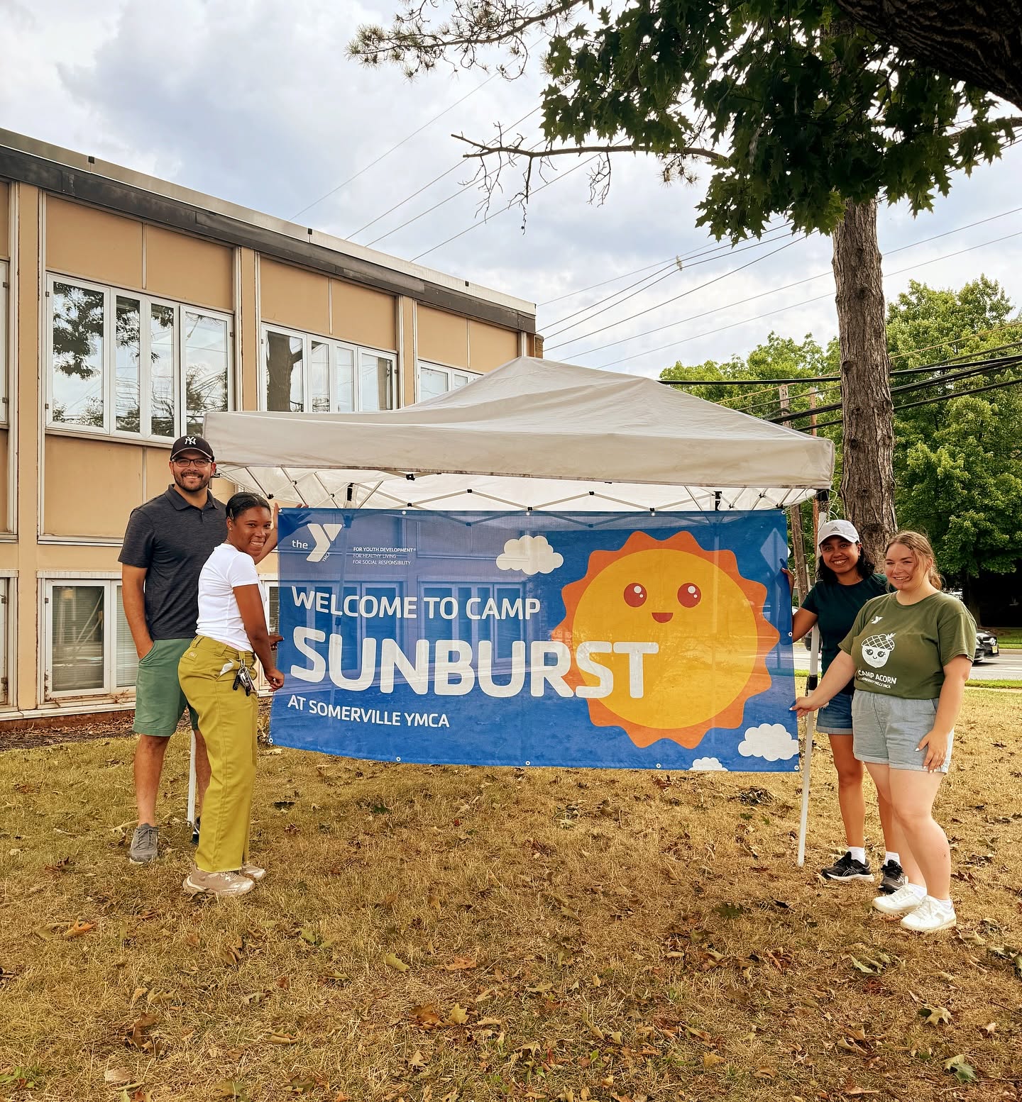 Four people standing outdoors near a tent and a banner that reads 'Welcome to Camp Sunburst' at the YMCA.