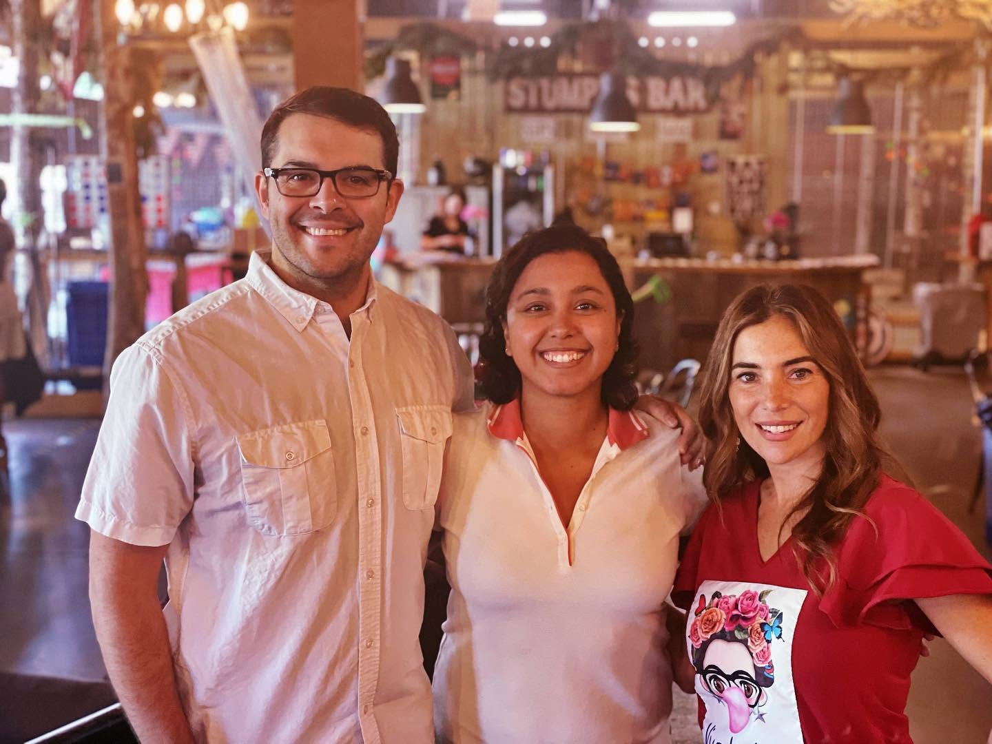 Three smiling people standing together indoors, with a blurred background of a market or food stall area.