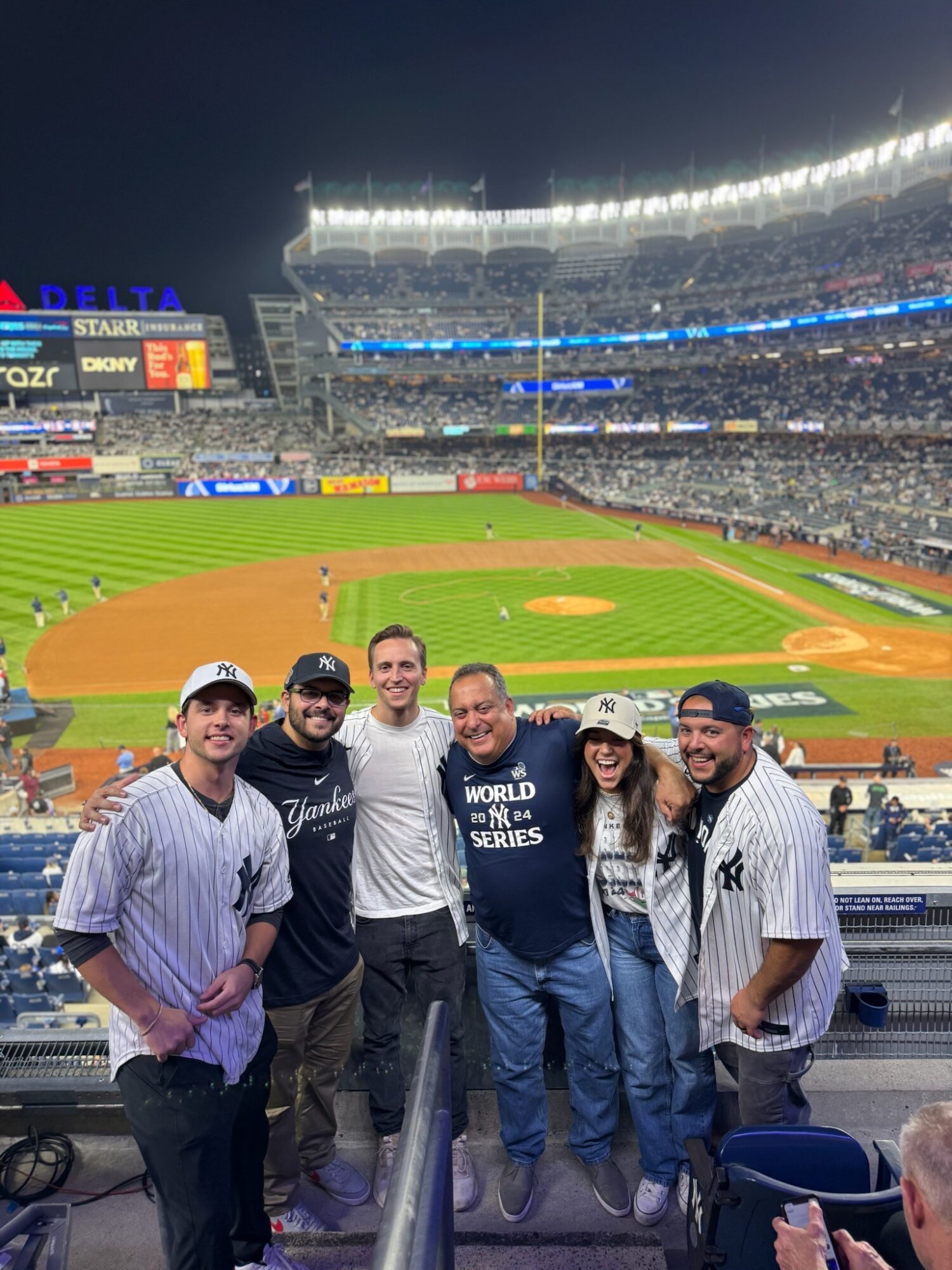 Group of six people smiling at a baseball stadium, with the field and stands in the background.