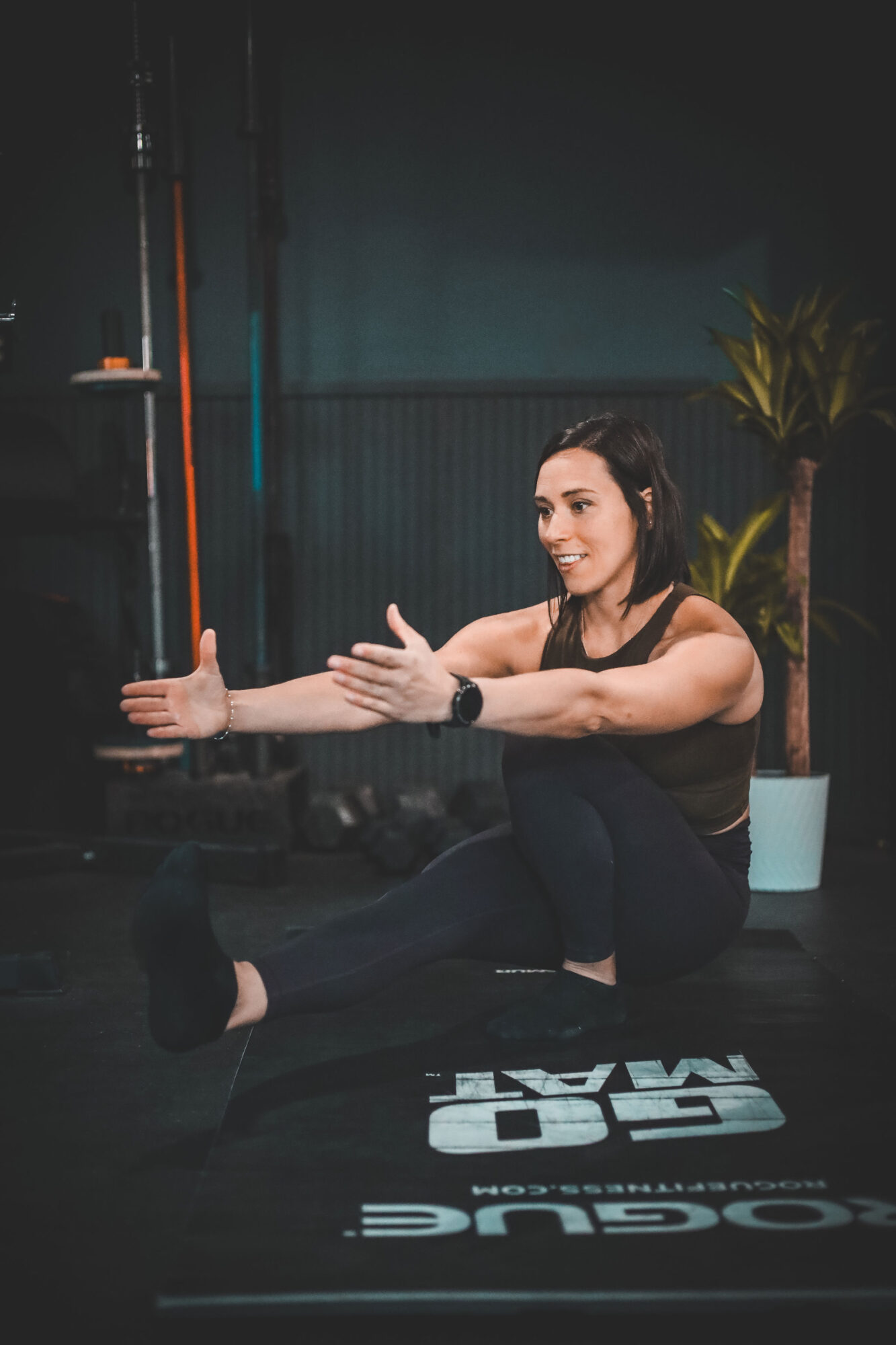 Woman sitting on a mat stretching with arms extended, smiling, in a gym with plants and equipment in background.