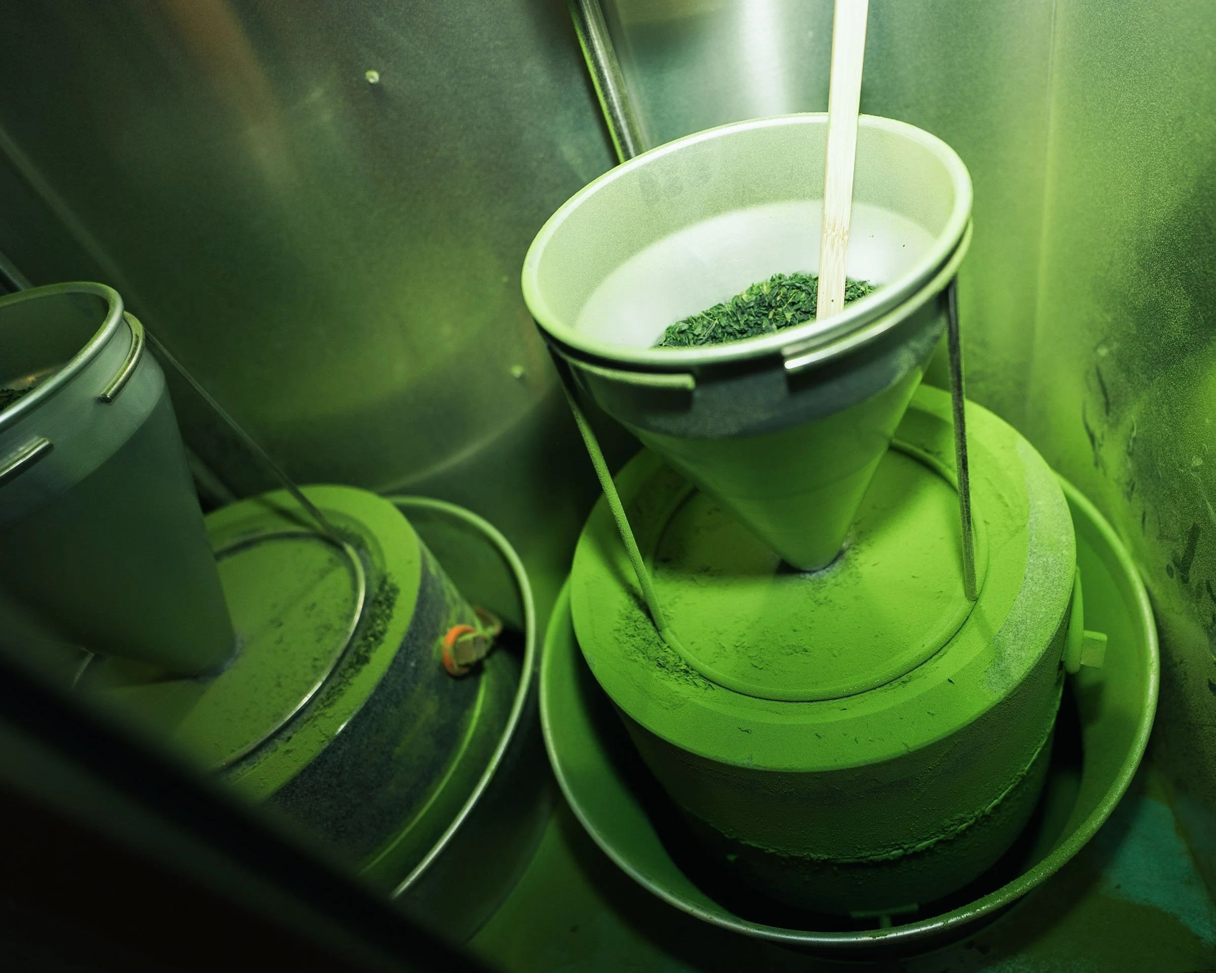 A green-colored machine with a funnel and a stick inside, surrounded by metallic equipment, in a green-lit environment.