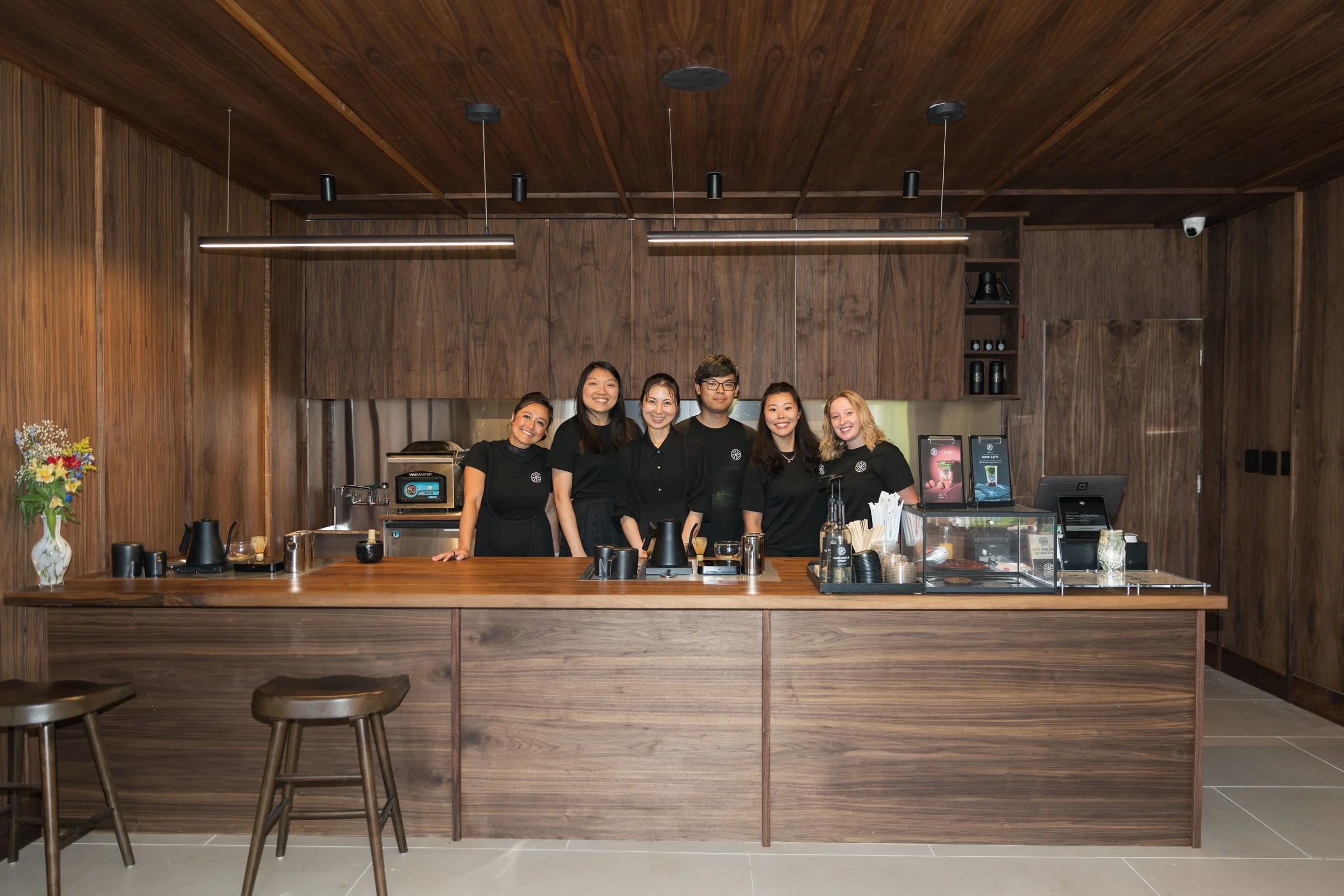 Six people stand behind a wooden bar counter in a dimly lit room with wood-paneled walls, smiling at the camera.
