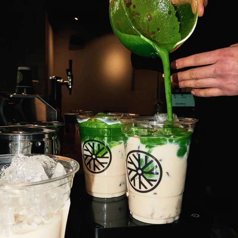 Person pouring green matcha latte into a cup with a logo, with other cups and a glass of ice nearby.