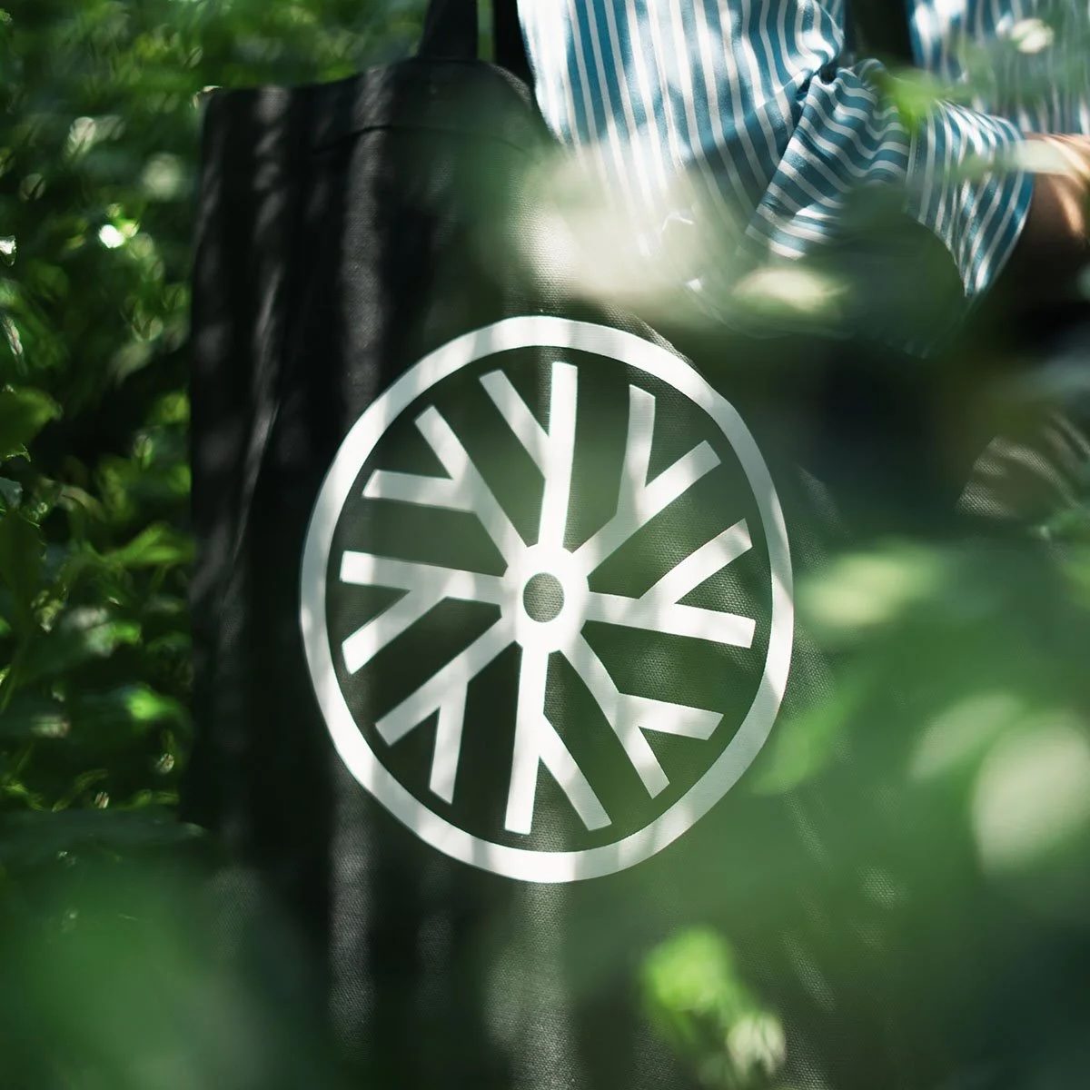 A black bag with a white circular logo, surrounded by green foliage and sunlight, with a person in a striped shirt nearby.