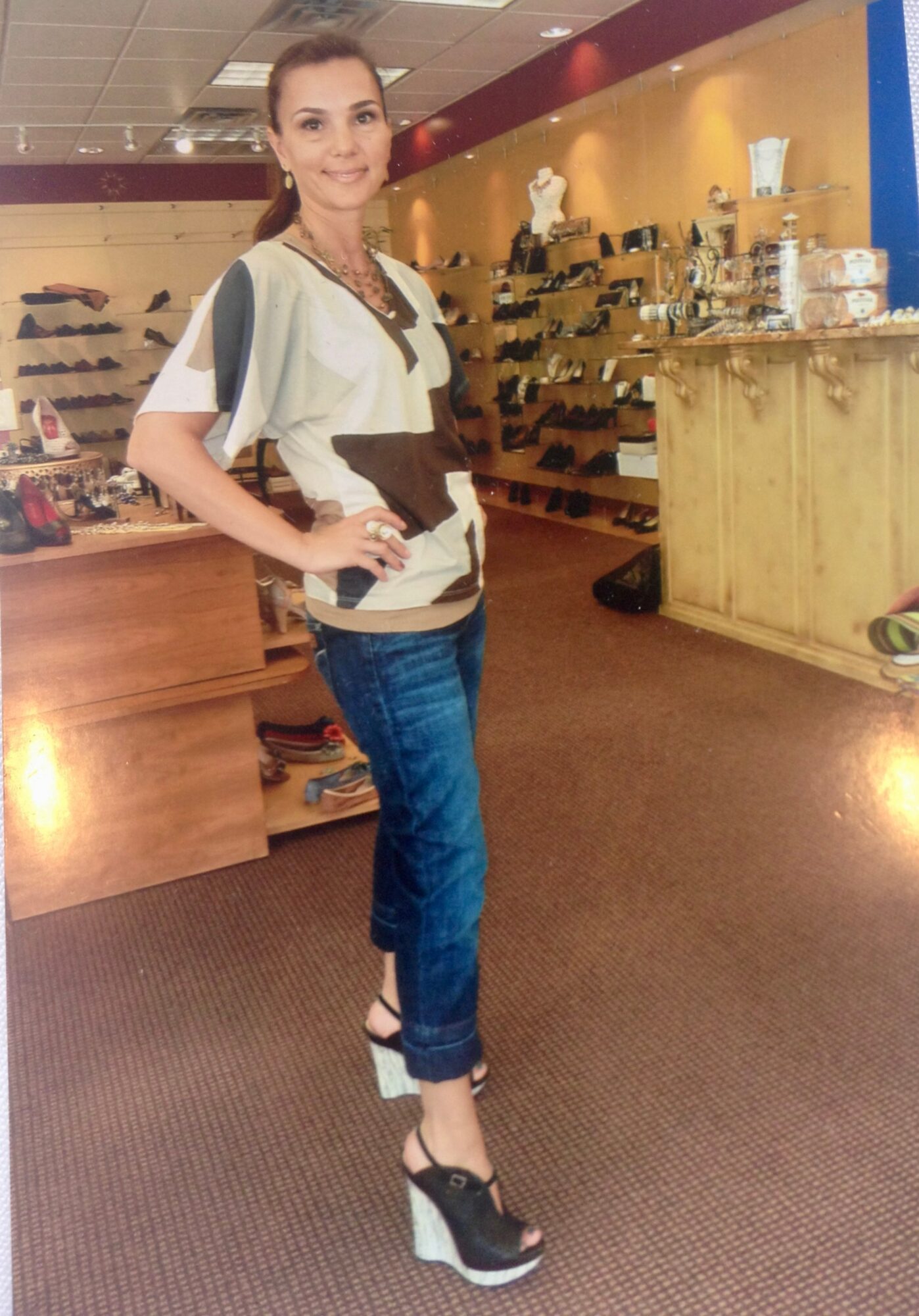 Woman standing in a store, smiling, wearing a beige and black top, jeans, and high-heeled shoes, with shelves and display in background.