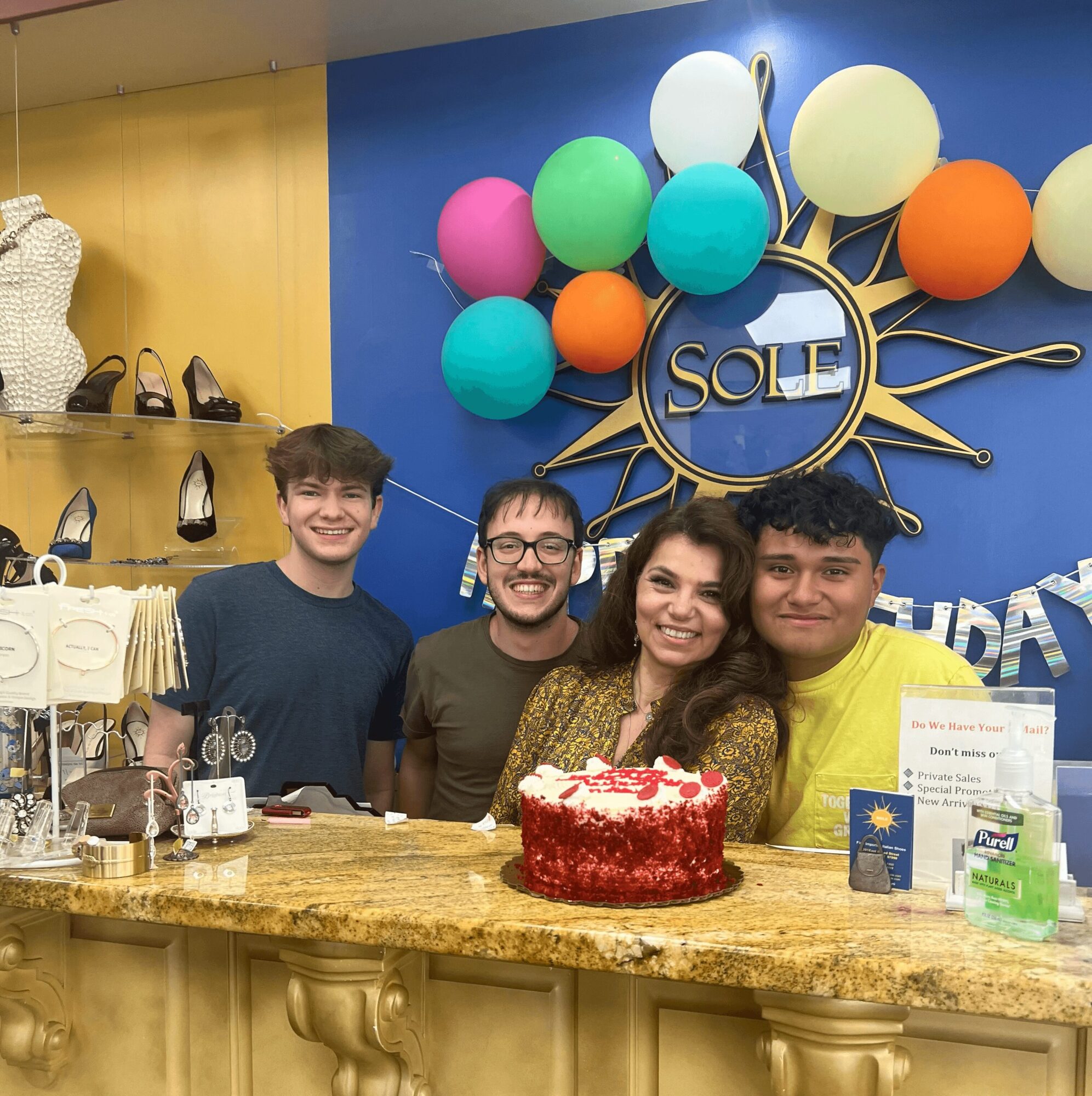 Four people smiling behind a cake on a store counter, with colorful balloons and a sign reading 'SOLE' in the background.
