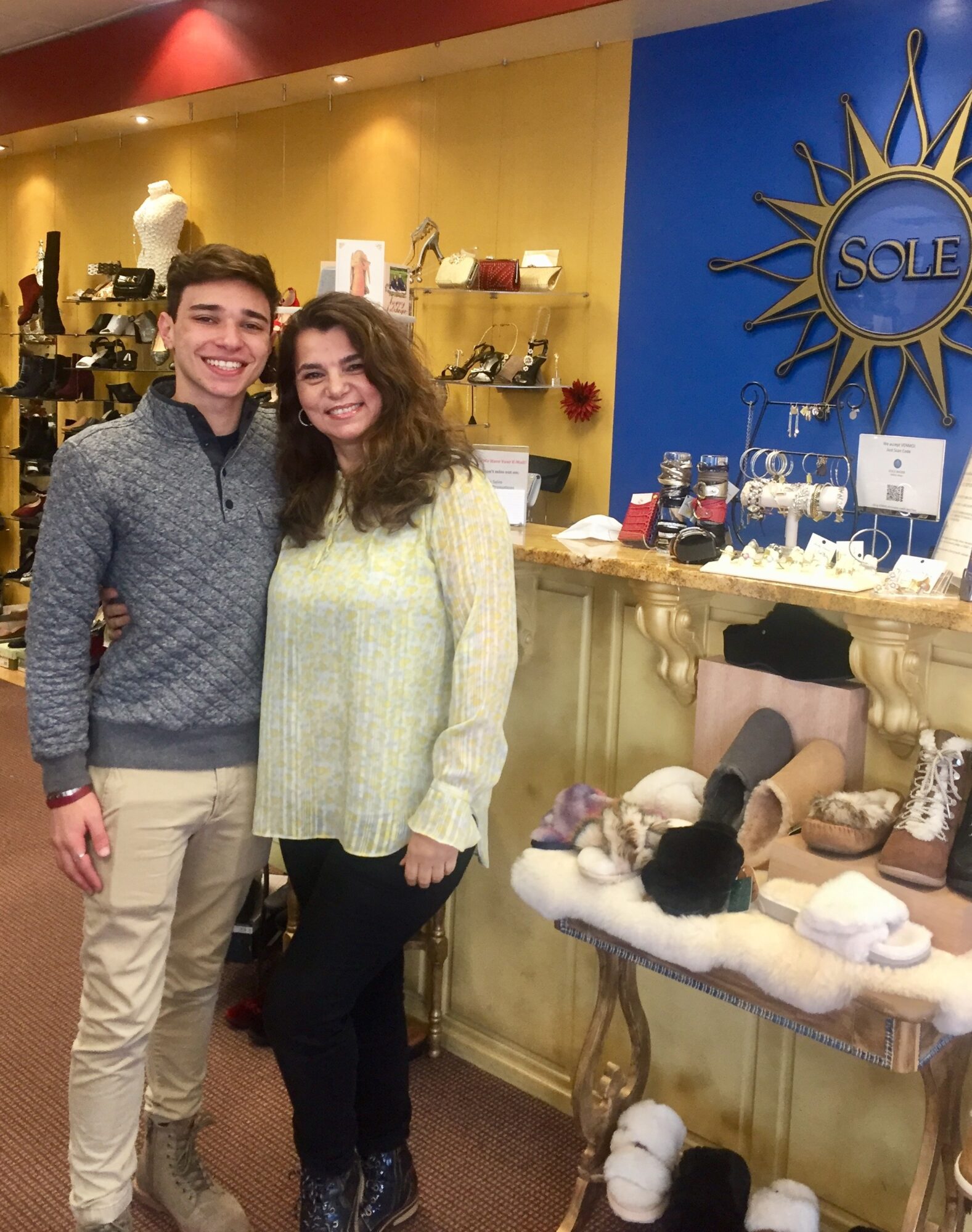 Two people standing inside a store, smiling, with shelves and jewelry displays behind them.