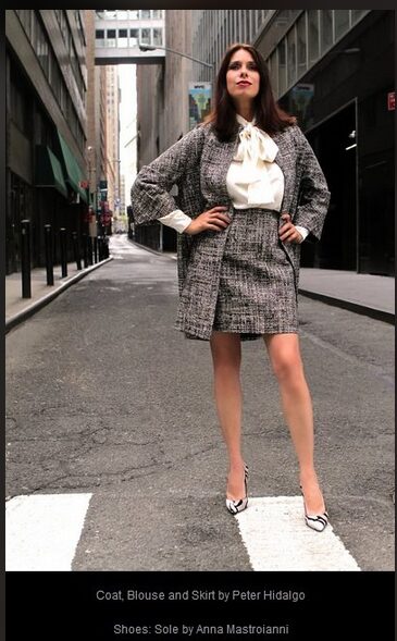 Woman standing confidently in city street wearing a coat, blouse, and skirt, with hands on hips and tall heels.